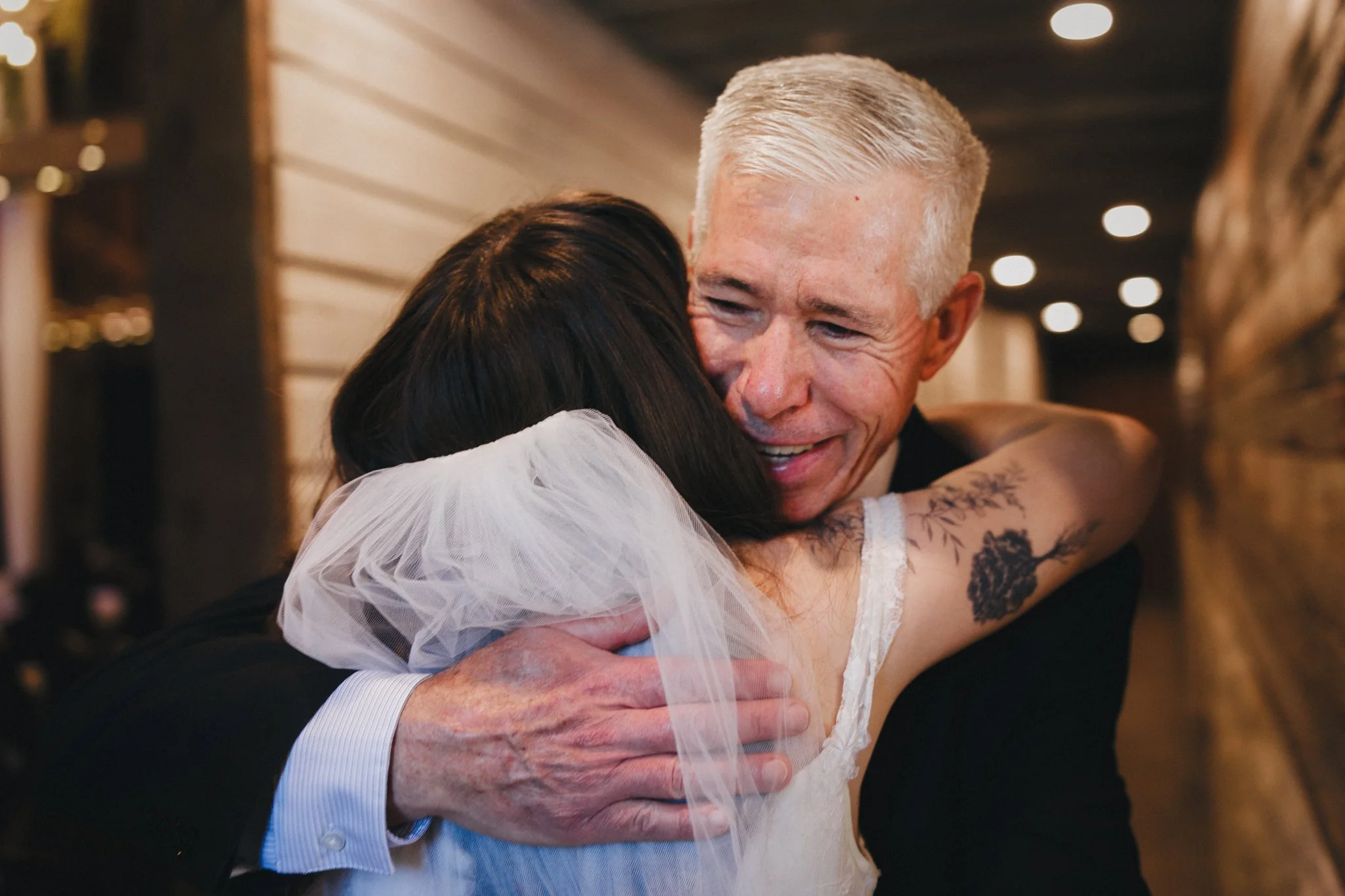 An elderly man with short white hair hugging a woman with dark hair during a wedding celebration.