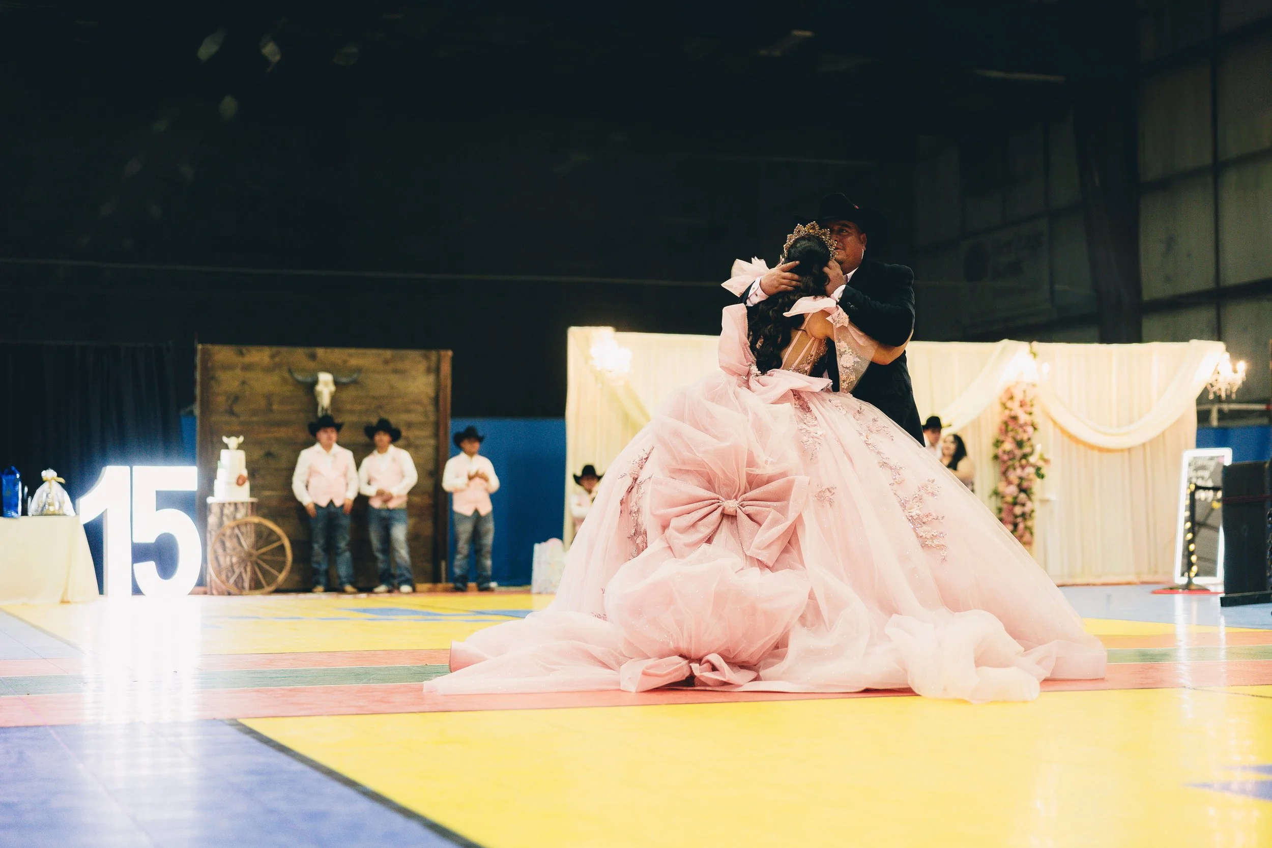 A young girl in a pink gown and a man in a cowboy hat dancing at a wedding reception. The girl’s dress has large bows. In the background, there are men in cowboy hats and a decorated wall with floral arrangements.