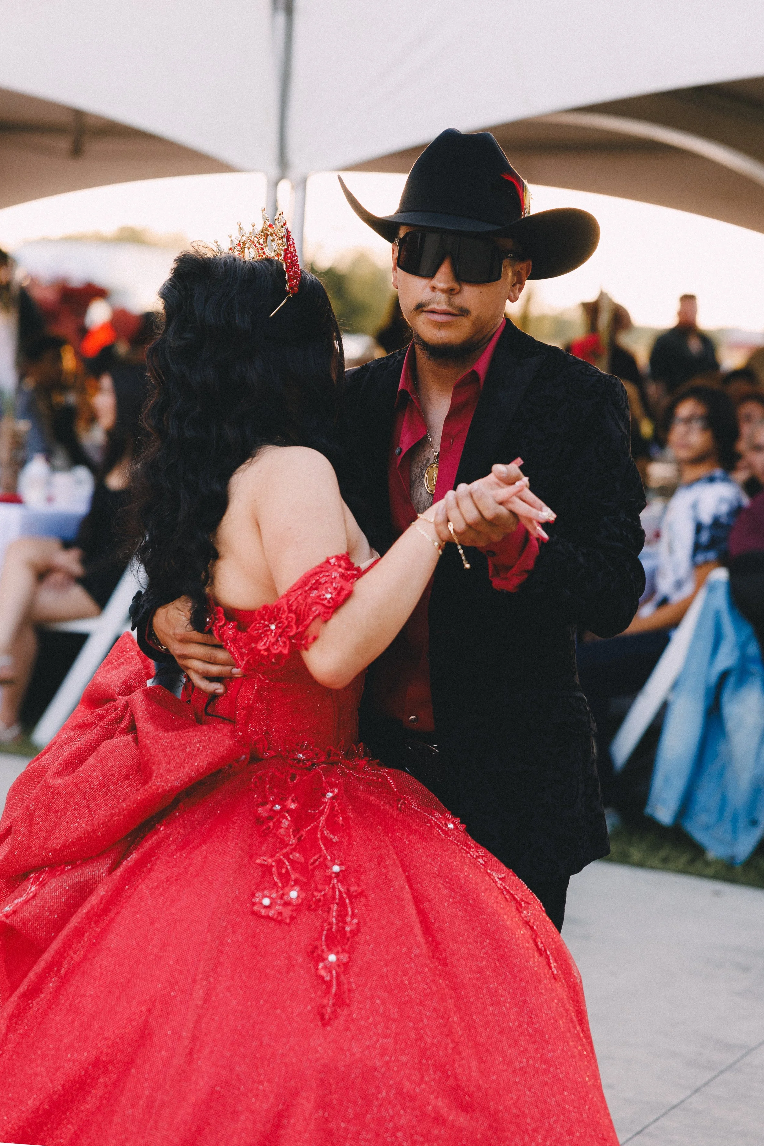 A couple dancing at an outdoor event, with the woman in a red gown and tiara, and the man wearing a cowboy hat, sunglasses, and a blazer.