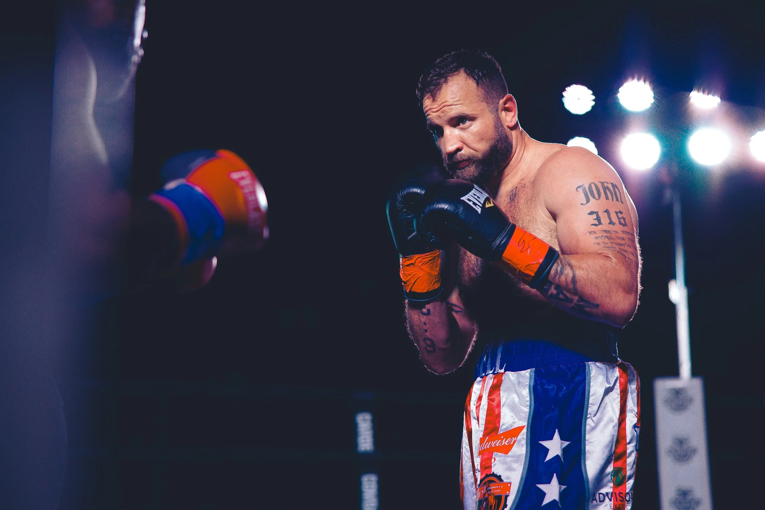 A male boxer with tattoos on his left arm wearing black gloves and colorful shorts, practicing boxing techniques in a dimly lit boxing ring with bright lights in the background. Shot at Evergy Plaza in Topeka, Kansas. 