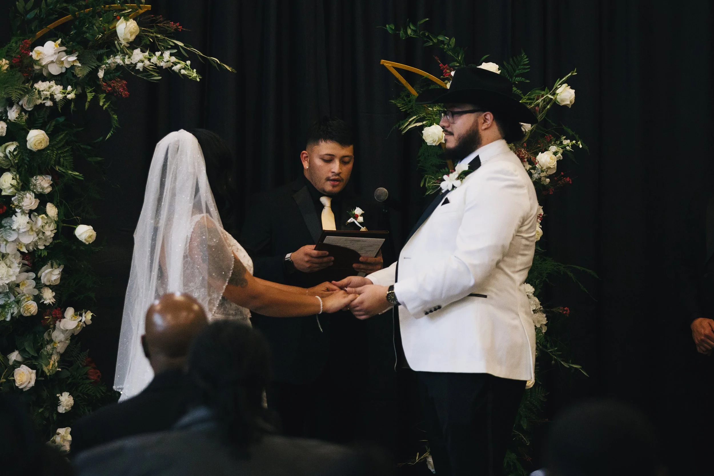 A wedding ceremony with a same-sex couple exchanging vows, one dressed in a white tuxedo with a black hat and glasses, the other in a black dress and veil, with an officiant reading from a booklet, surrounded by floral arrangements and black curtains