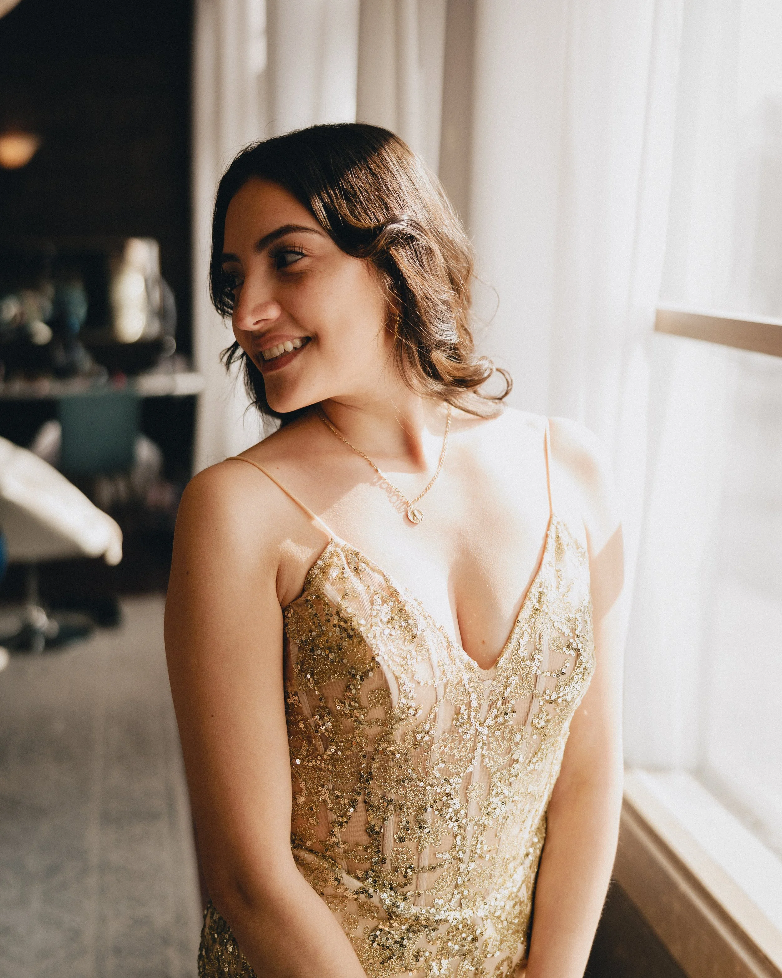 A young woman with dark brown hair and a smile, wearing a gold, sequined dress with thin straps, standing near a window with sheer curtains, sunlight illuminating her face. Shot at Topeka ,Kansas venue The Beacon.