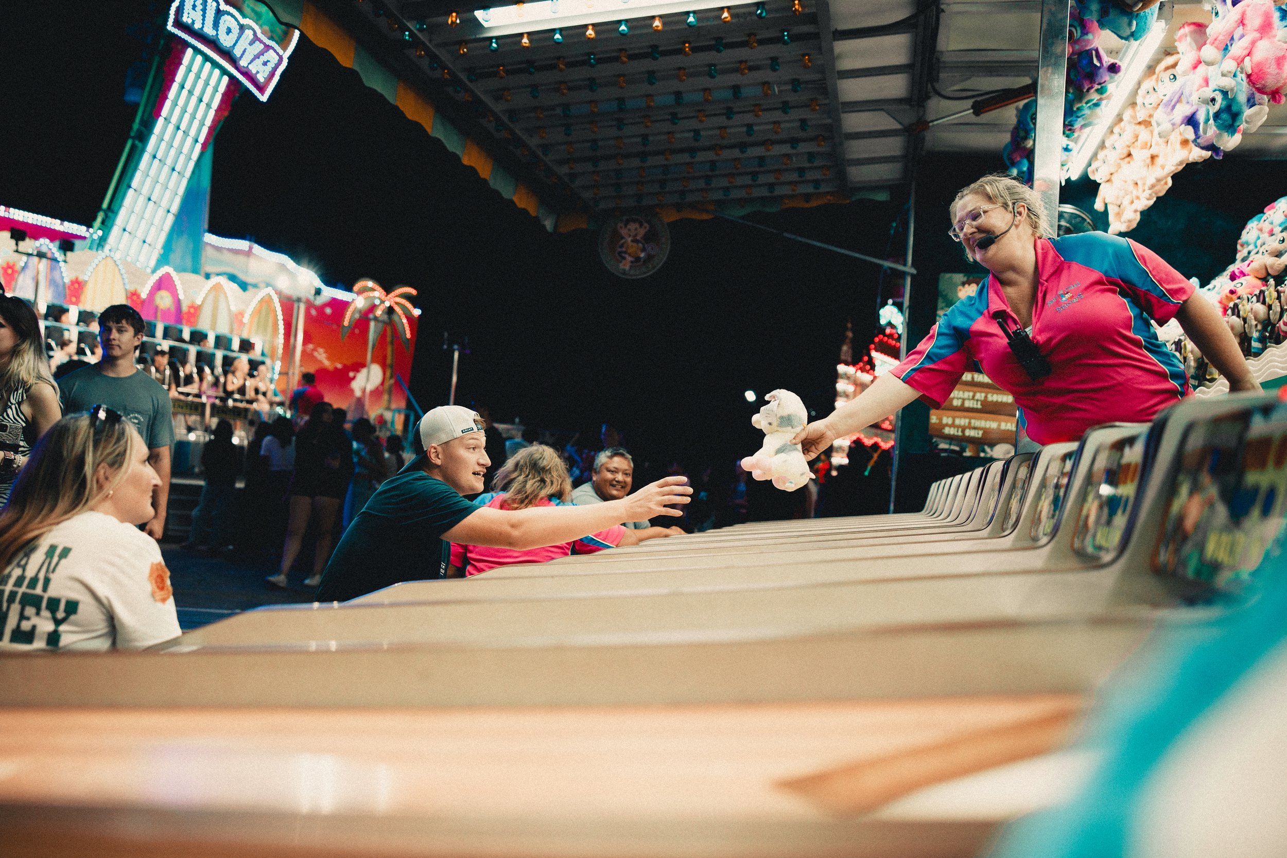 A girl at a carnival game extends her hand to give a stuffed unicorn toy to a young man sitting at the game booth. Several people in the background are enjoying the carnival rides and attractions under bright, colorful lights at night. Shot at Fiesta