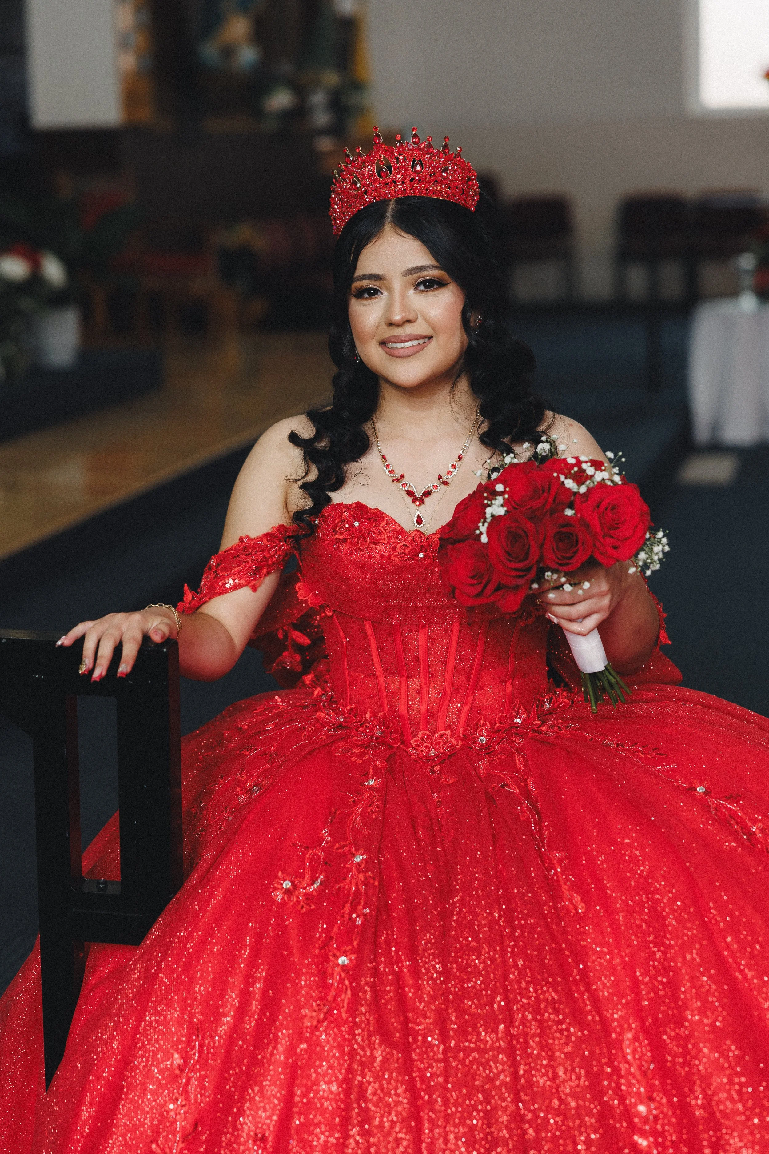 Young woman dressed in a red gown, wearing a crown, holding a bouquet of red roses, and smiling at the camera.