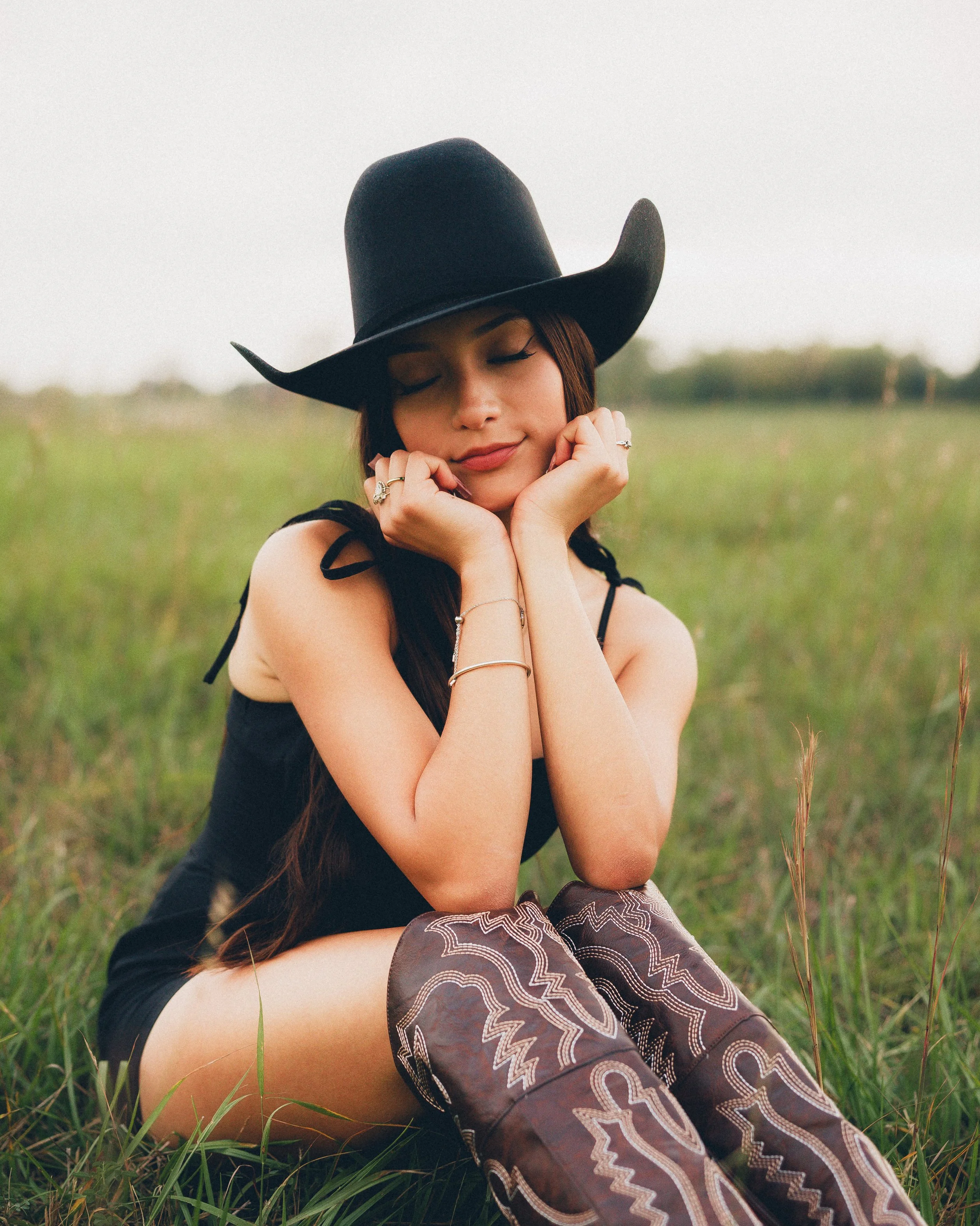 A young woman with long brown hair, wearing a black wide-brimmed hat, a black sleeveless dress, and patterned chaps, sitting in a grassy field with her eyes closed and a serene expression.