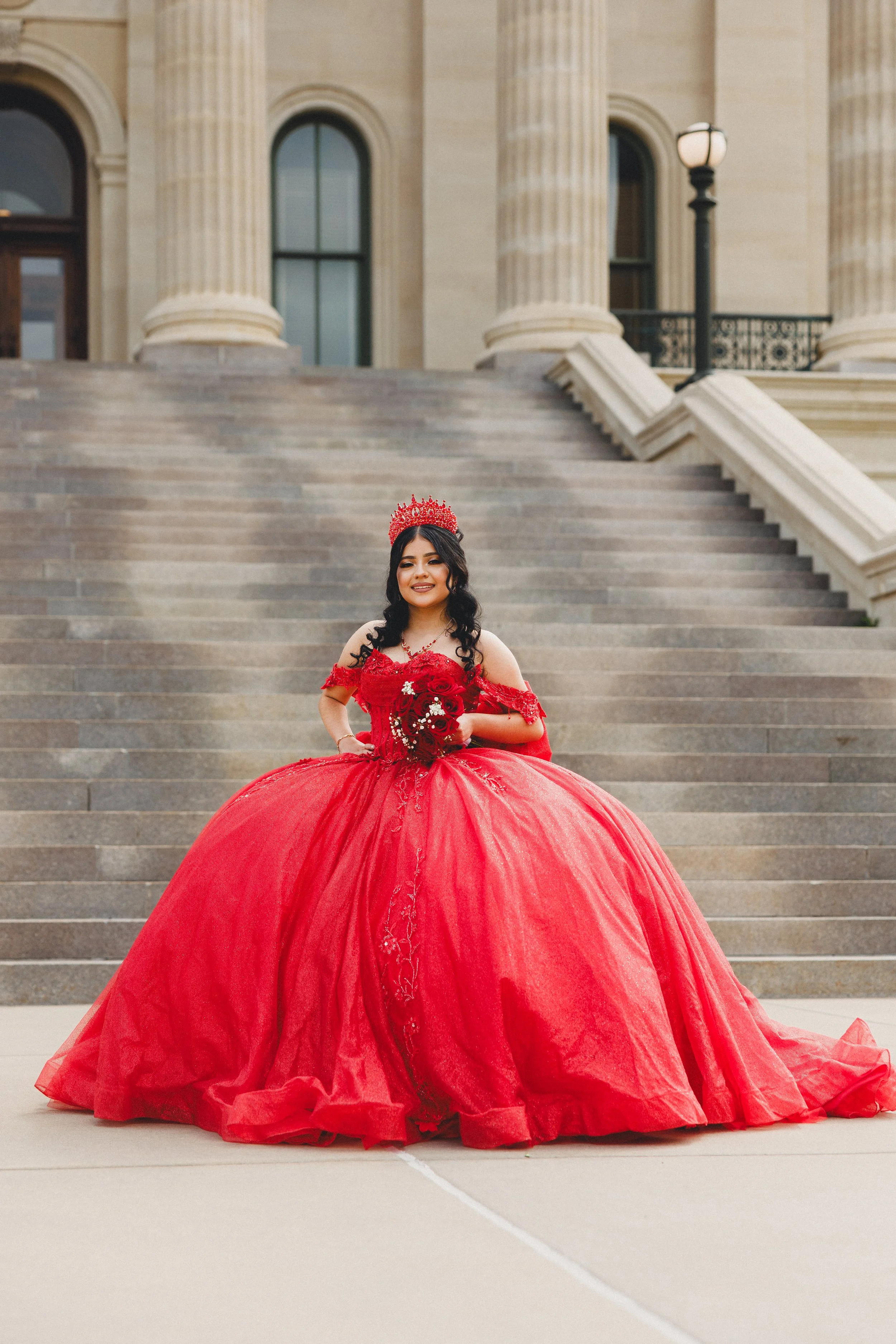 Young woman in a red ball gown with lace details, wearing a matching crown and holding a bouquet, standing on steps in front of a historic building with columns and a lamp post.