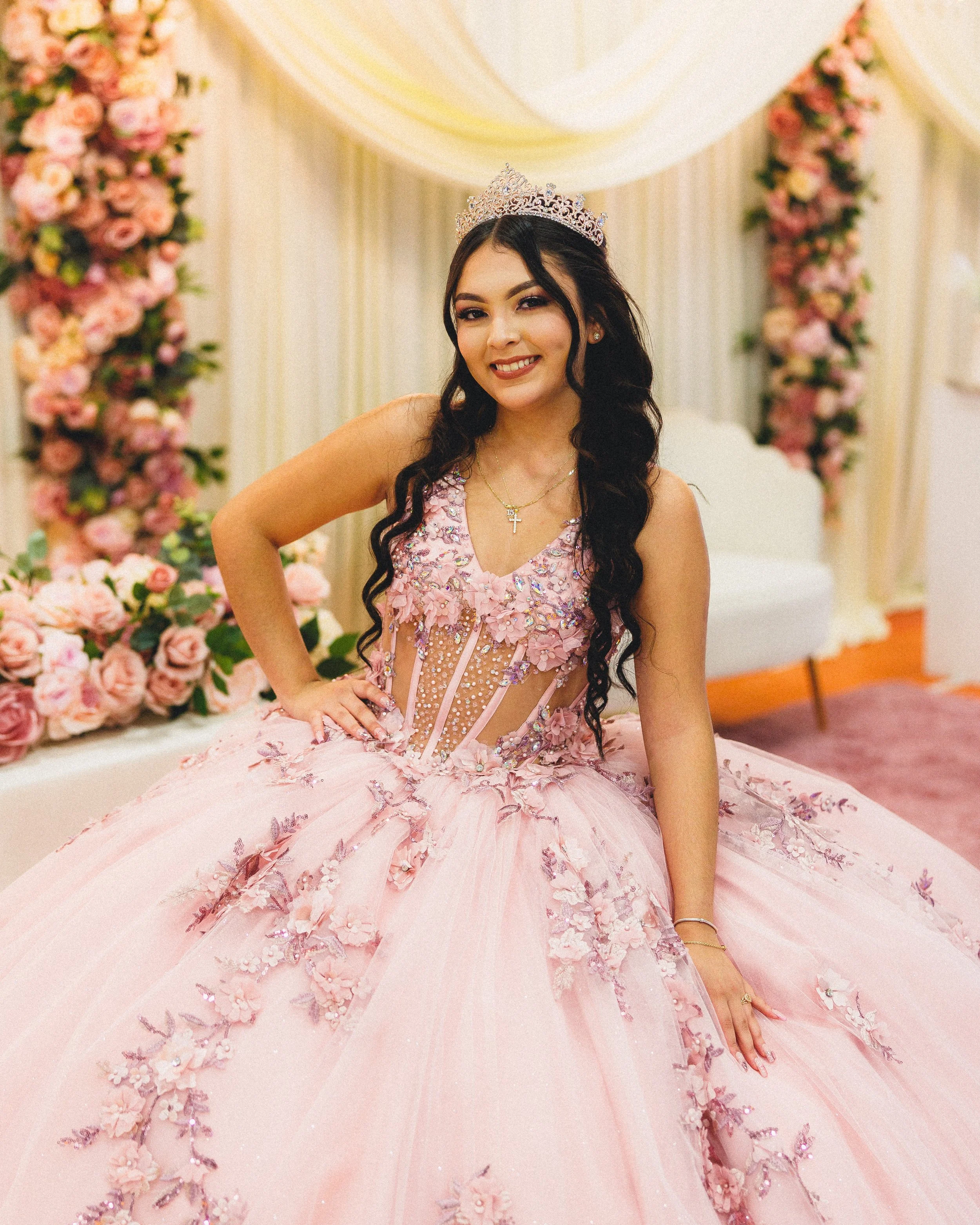 Young woman in a pink quinceañera dress and tiara, smiling, in a decorated room with pink and peach flowers and a white backdrop.