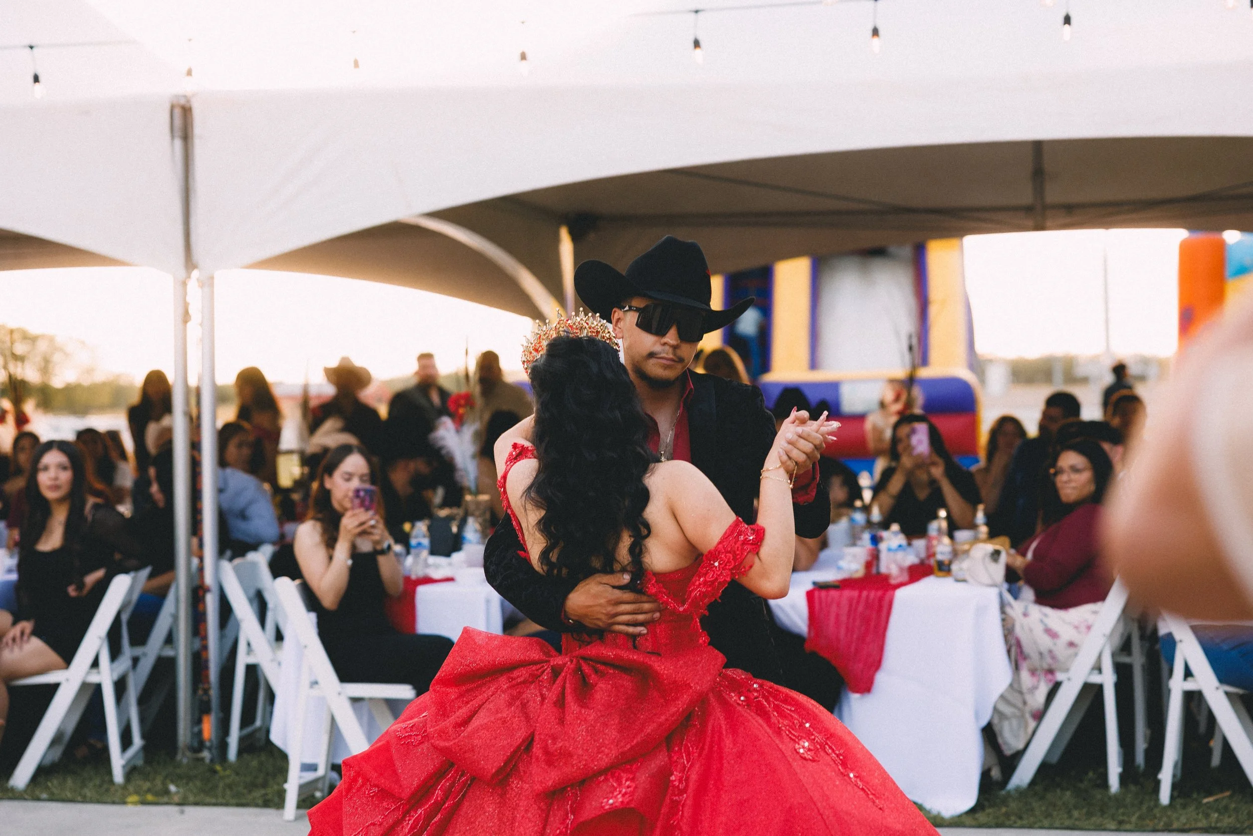 A couple dancing at an outdoor event under a large white canopy with guests seated at tables in the background.