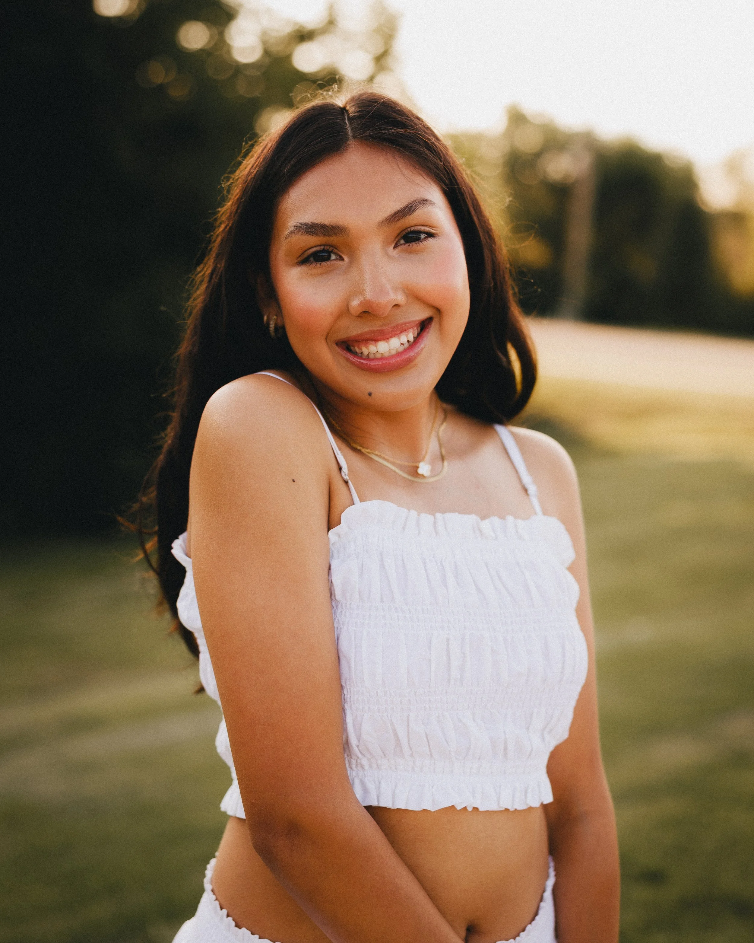 A young woman with long dark hair, wearing a white sleeveless top and gold jewelry, smiling outdoors during sunset.