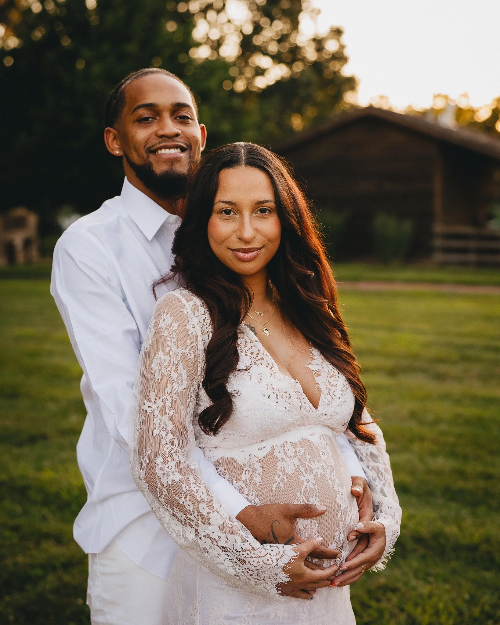 A happy couple, with the man standing behind the woman, both smiling, at sunset outdoors. The woman is pregnant, holding her belly, and the man has his arms around her. Shot in Topeka, Kansas. 