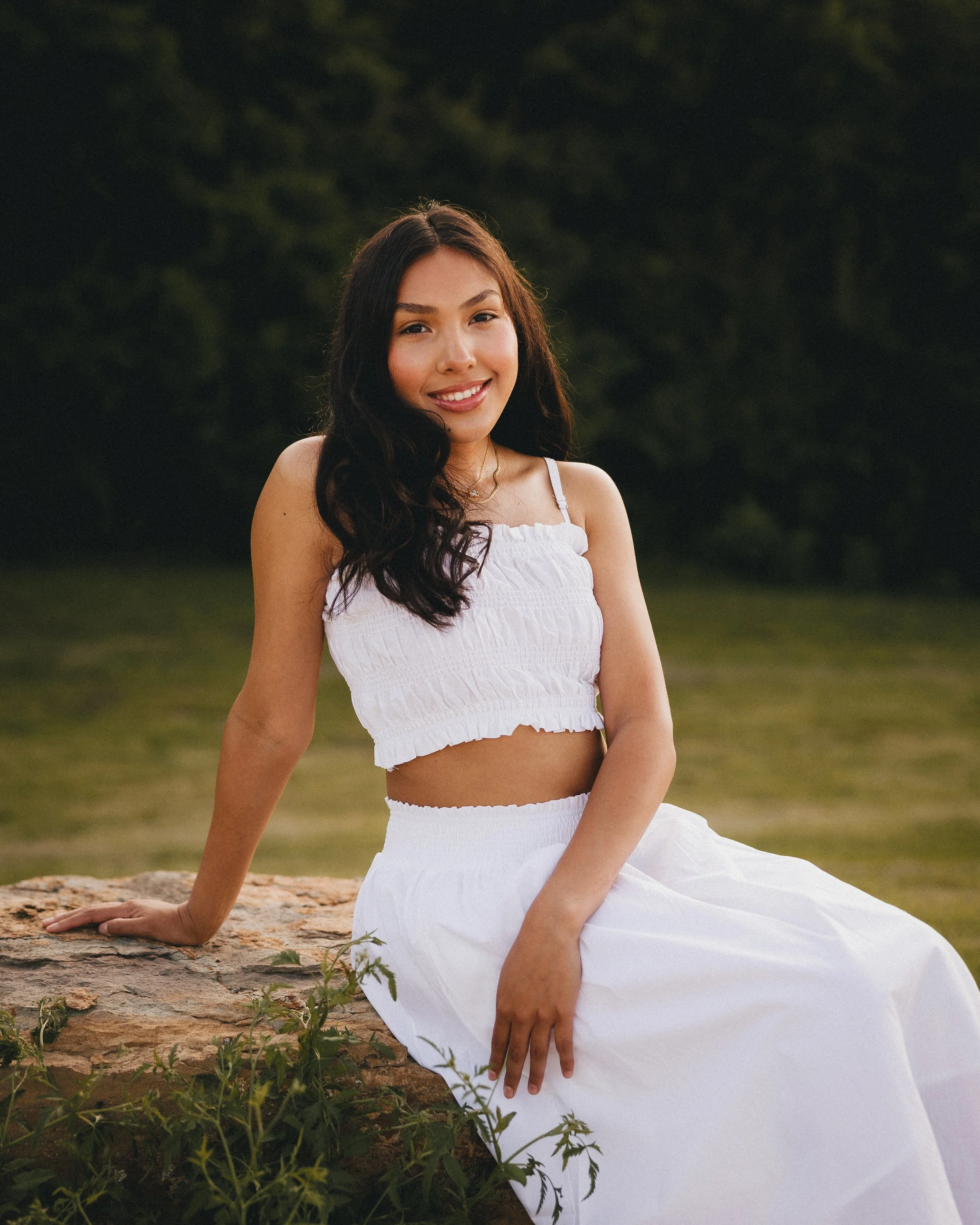 A young woman with long dark hair and light skin is sitting on a rock outdoors, smiling at the camera, wearing a white cropped top and long white skirt with a dark green background.