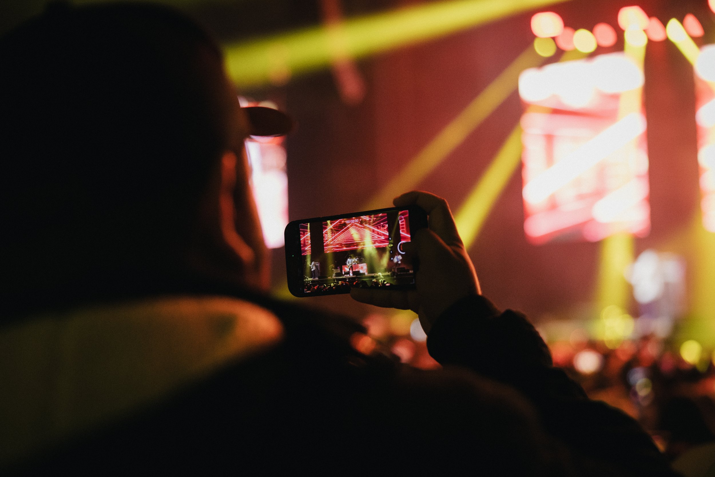 Person taking a photo or recording a video of a concert or show on a smartphone, with colorful stage lights and screens in the background. Los Tucanes de Tijuana Stormont Vail Event Center Topeka, Kansas. 