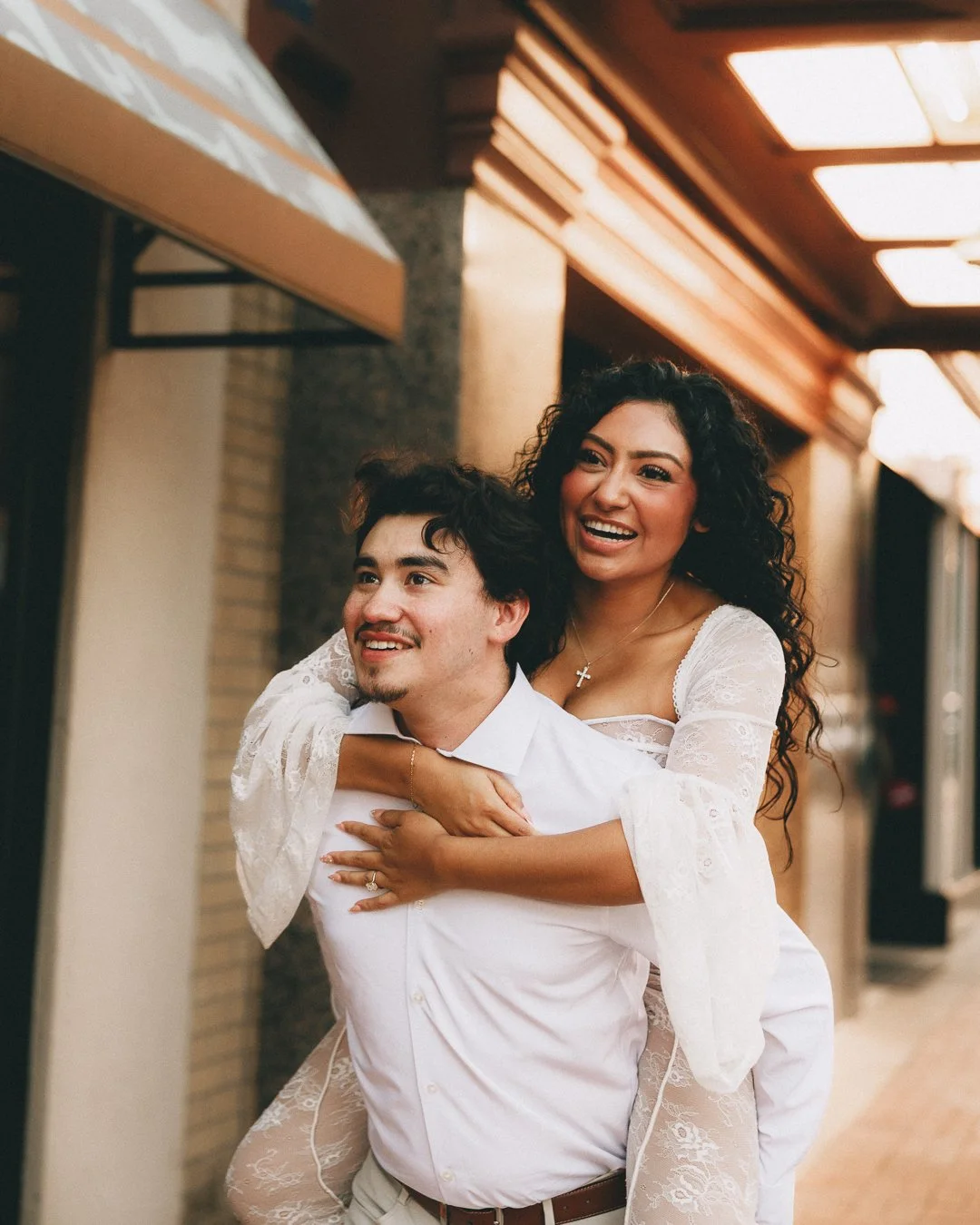 A woman with curly black hair wearing a white dress is on the man's back, smiling and hugging him, while the man with dark hair and a beard is wearing a white shirt. Shot at Kansas City, Country Club Plaza.