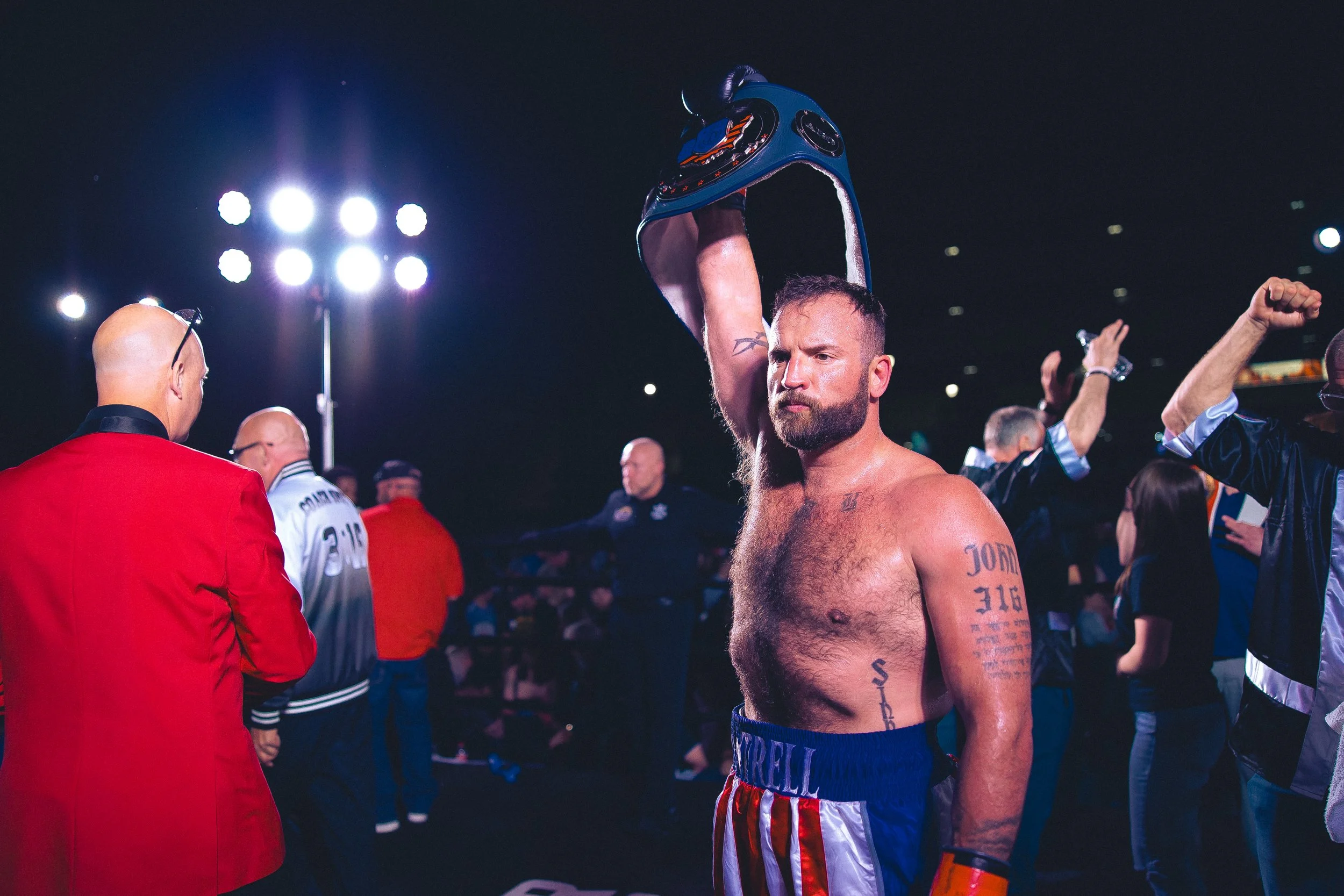 Topeka, Kansas boxer John Cantrell seen with tattoos and a beard, wearing boxing shorts and gloves, holding a boxing helmet above his head, standing in a boxing ring surrounded by people, under the bright open-air Evergy Plaza lights at night.