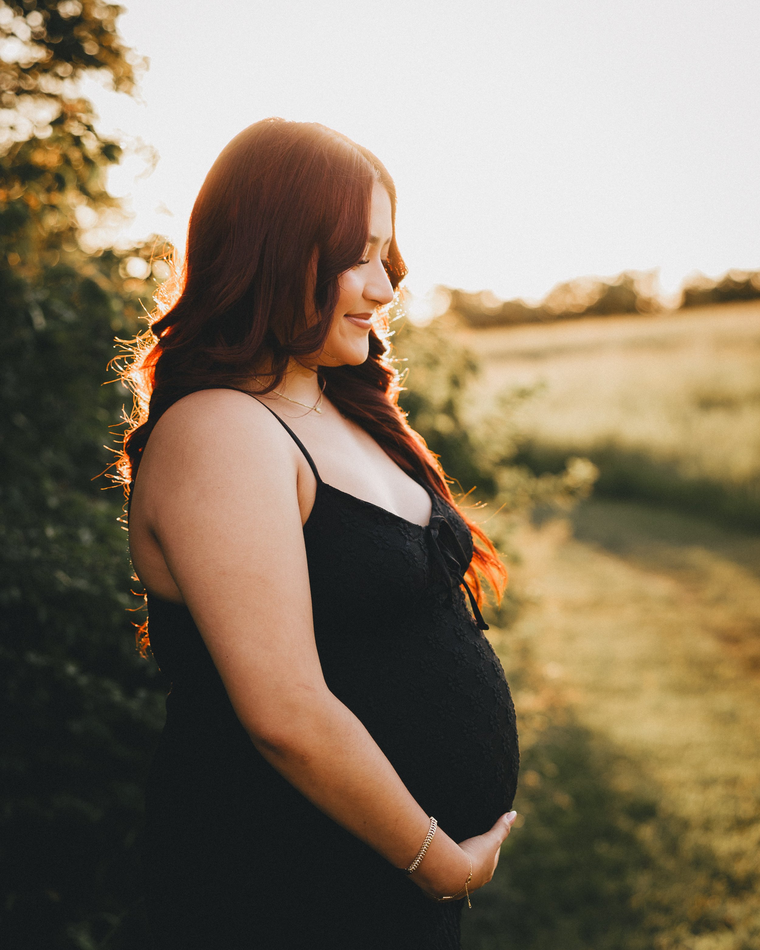 A pregnant woman with long red hair in a black dress standing outdoors during sunset, holding her belly with one hand.  Shot in Topeka, Kansas. 