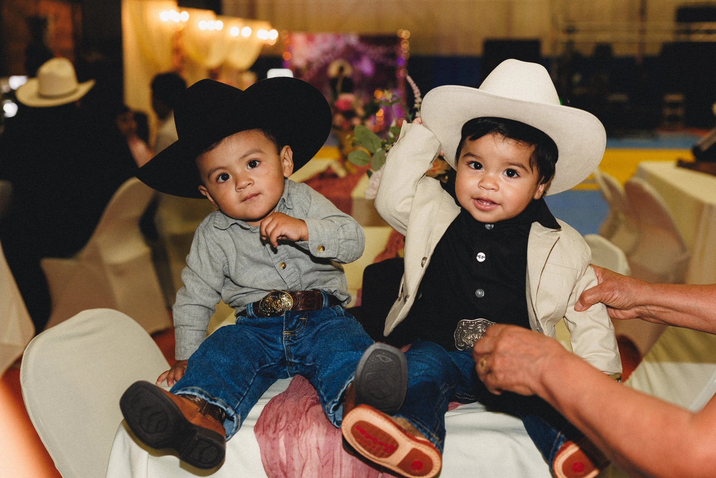 Two young children wearing cowboy hats, sitting on a table at a festive event. The child on the left wears a black cowboy hat, a gray shirt, jeans, and boots. The child on the right wears a white cowboy hat, a beige vest, dark shirt, and jeans. They 