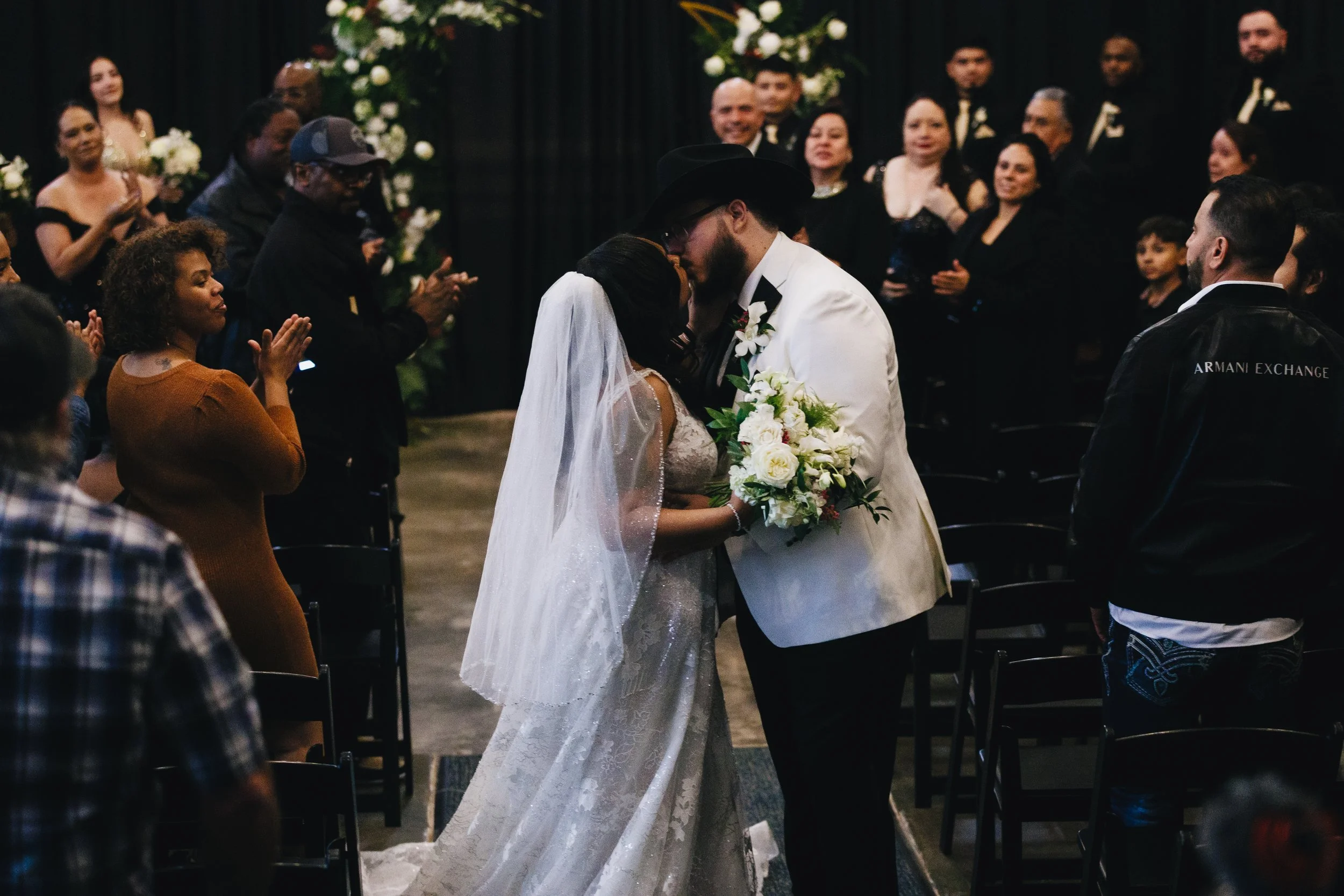 Couple exchanging wedding vows in a dimly lit indoor wedding ceremony, with guests standing around in front of a black curtain backdrop and floral decorations. Shot at Topeka Kansas venue The Beacon.
