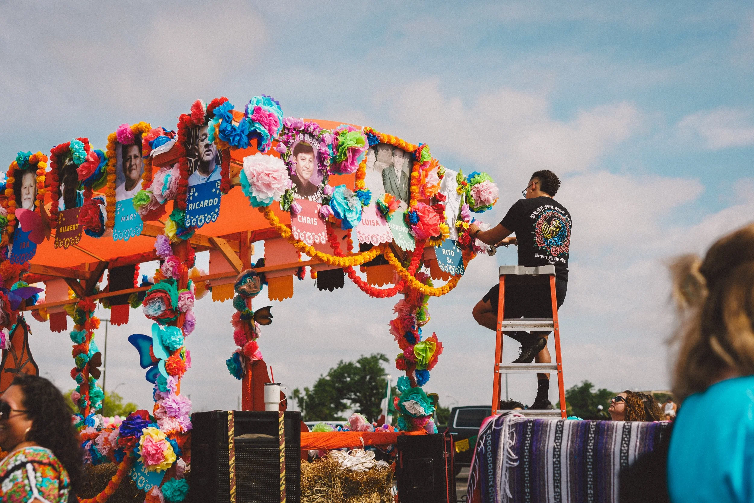 A colorful parade float decorated with paper flowers and photos of people, with a man on a ladder attaching decorations, under a partly cloudy sky. Shot at Fiesta Topeka parade.