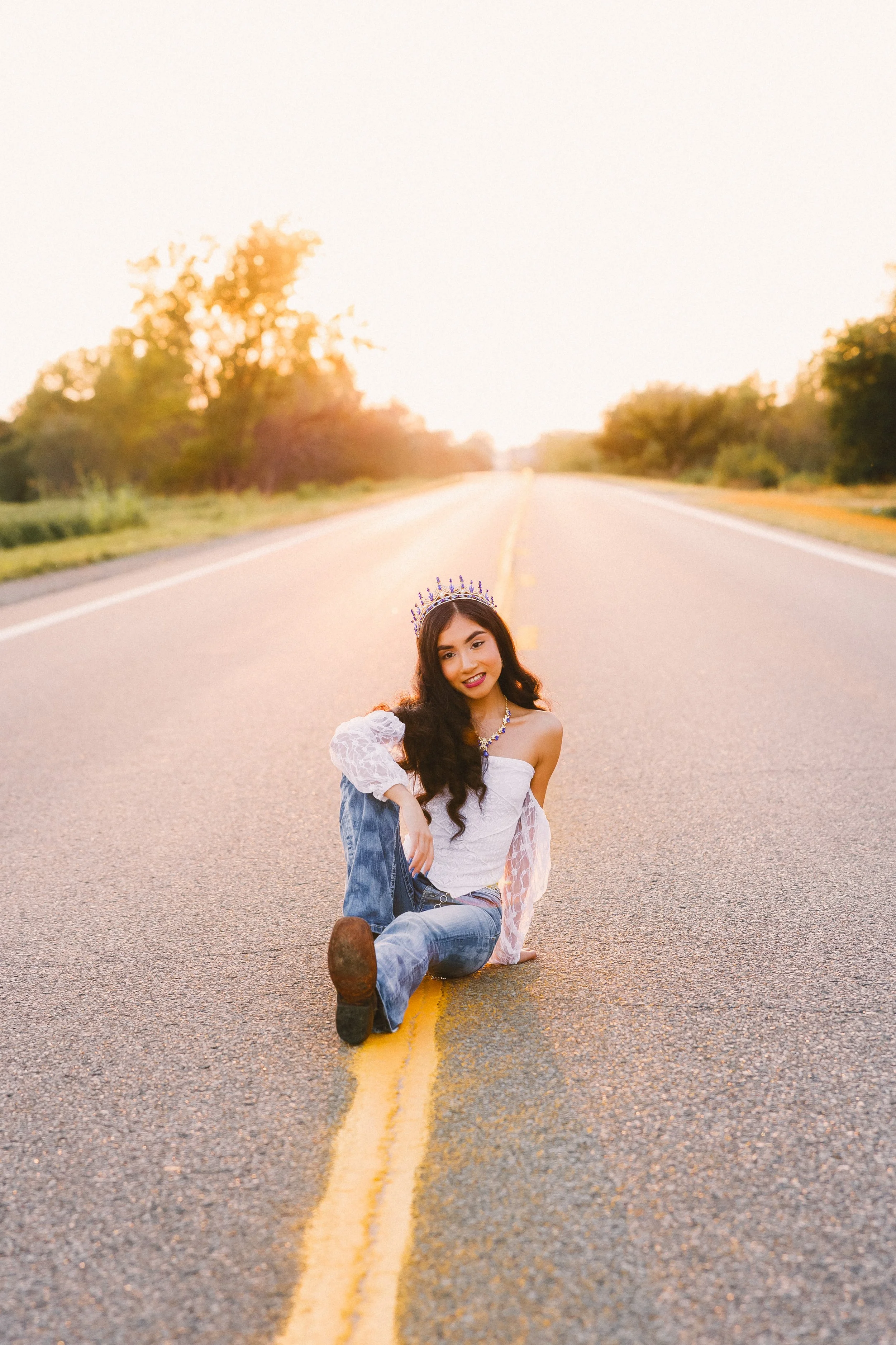 Young woman sitting on a rural road at sunset, wearing a white off-shoulder top, jeans, a crown, and jewelry, smiling at the camera.