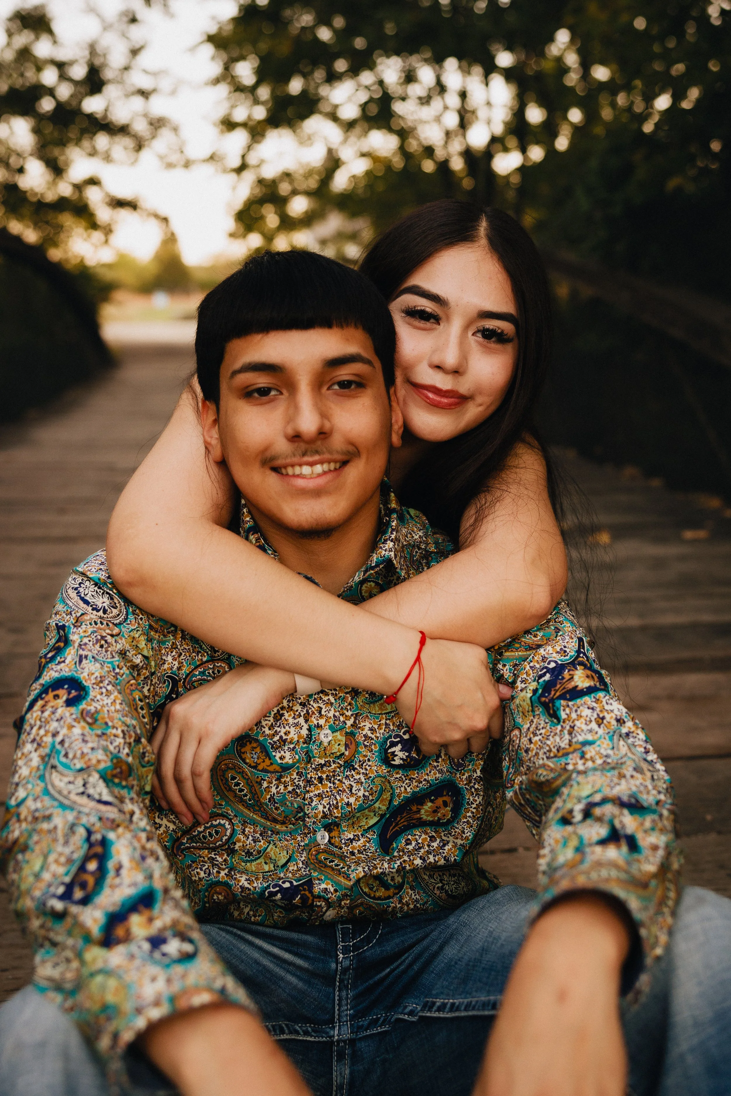 A young couple sitting on a wooden bridge outdoors during sunset, with the woman hugging the man from behind, both smiling at the camera. Shot in Topeka, Kansas. 