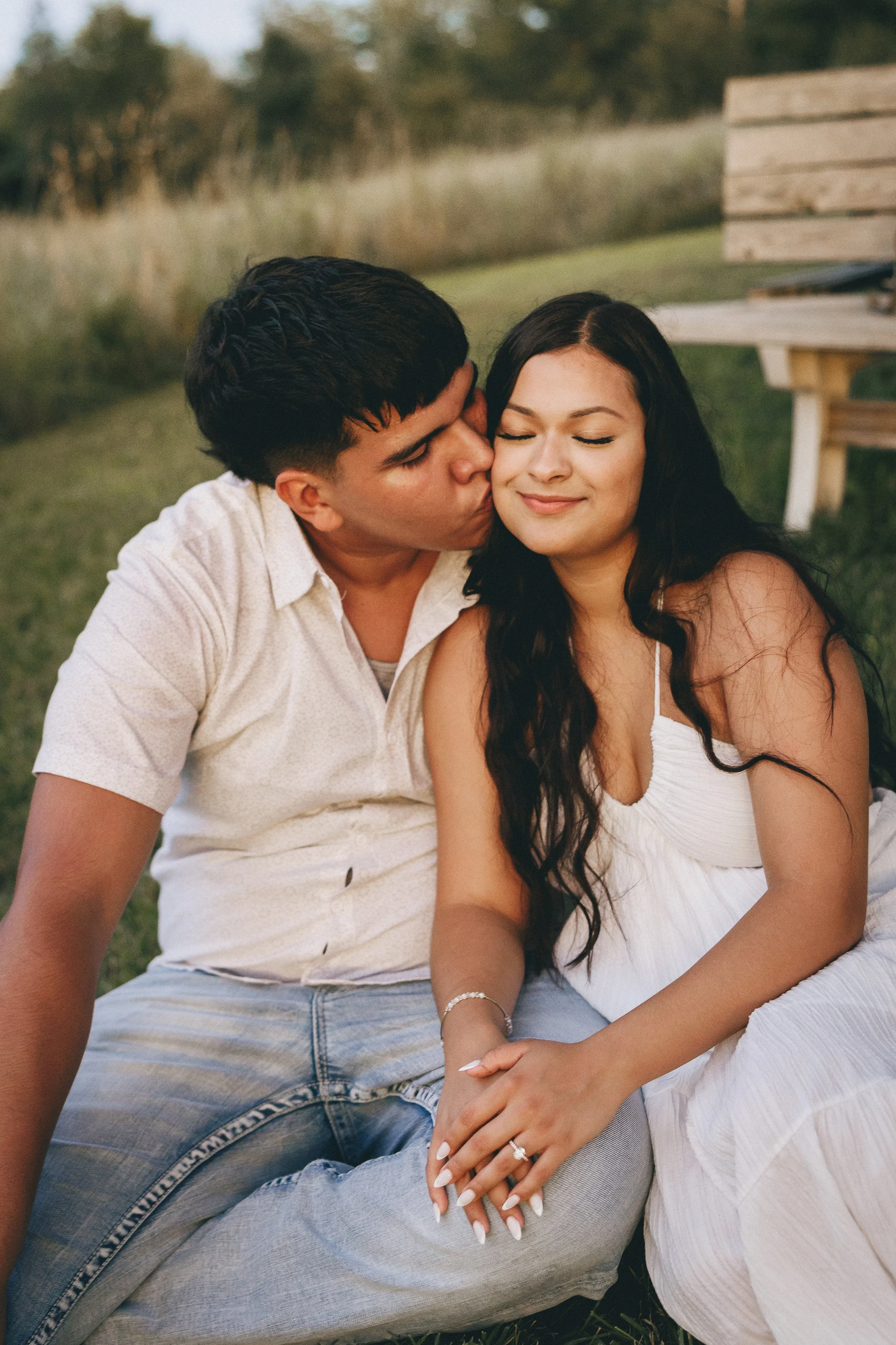 A young man and woman sitting on grass outdoors, the man kissing the woman on the cheek, she smiling with eyes closed, in a natural setting with trees and a wooden bench in the background.  Shot in Topeka, Kansas. 