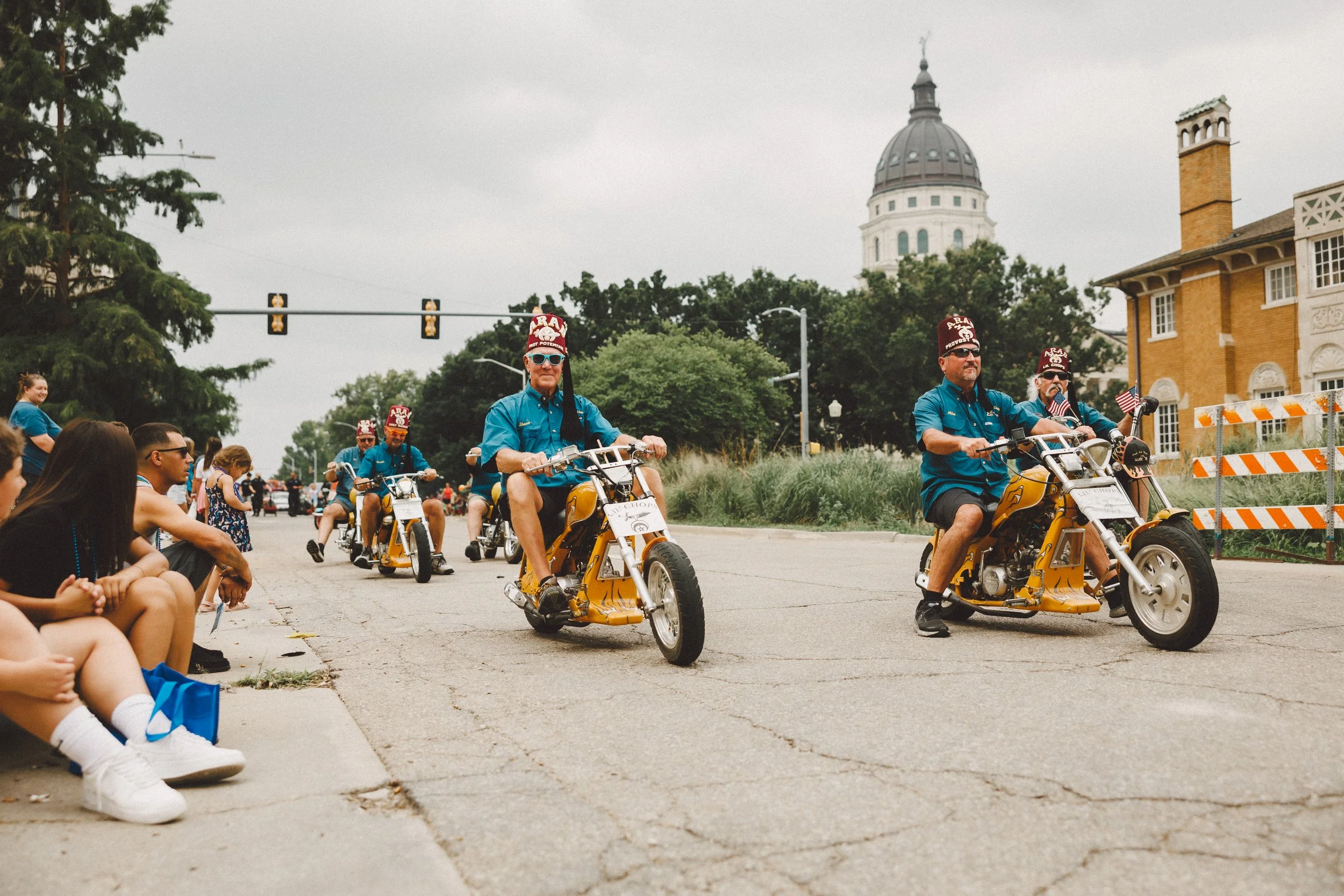 Group of Veterans riding motorcycles in a parade down a city street with spectators on the sidewalk, historic buildings, and a domed government building in the background. Shot at Fiesta Topeka parade.