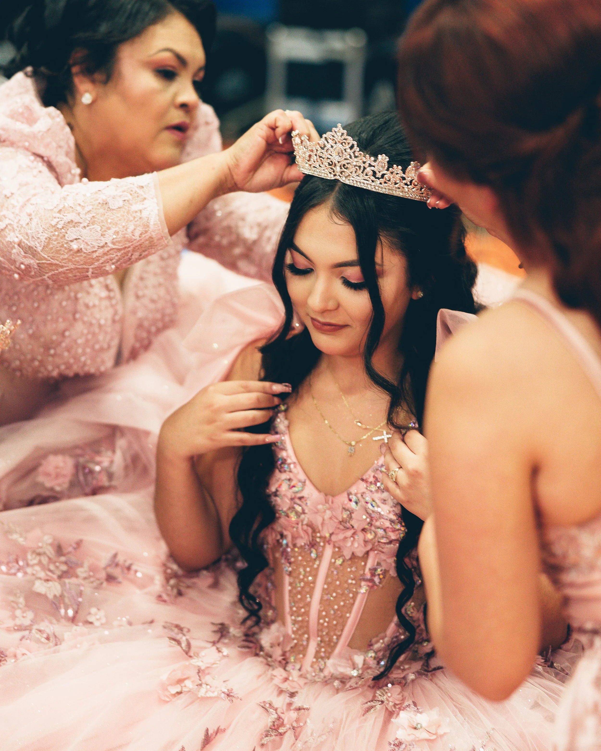 A young woman dressed in a pink, floral, and beaded gown is being crowned with a tiara by two women dressed similarly. The woman has long dark hair and a gentle expression.