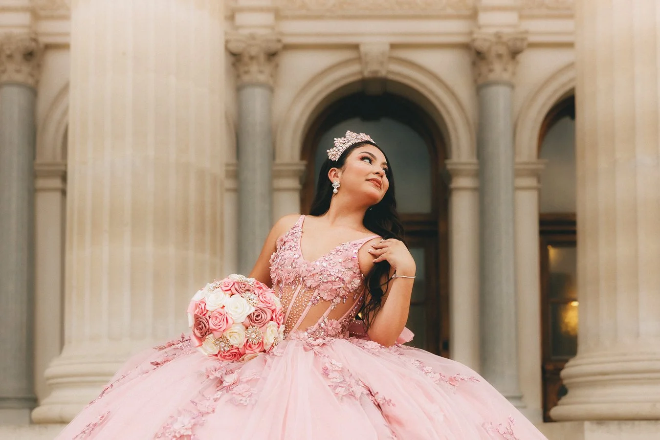 A woman dressed in a pink quinceañera dress with floral embellishments, holding a bouquet of pink and white roses, standing in front of grand marble columns and arches.