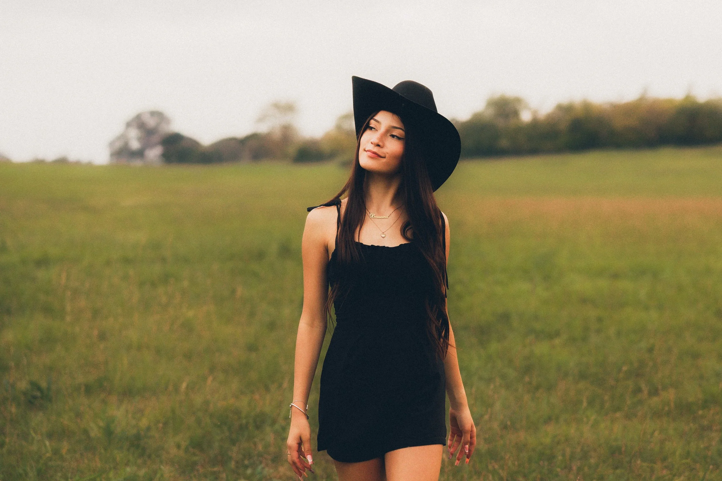 A young woman with long dark hair wearing a black dress and wide-brimmed black hat, standing in an open grassy field with trees in the background, during sunset or late afternoon.