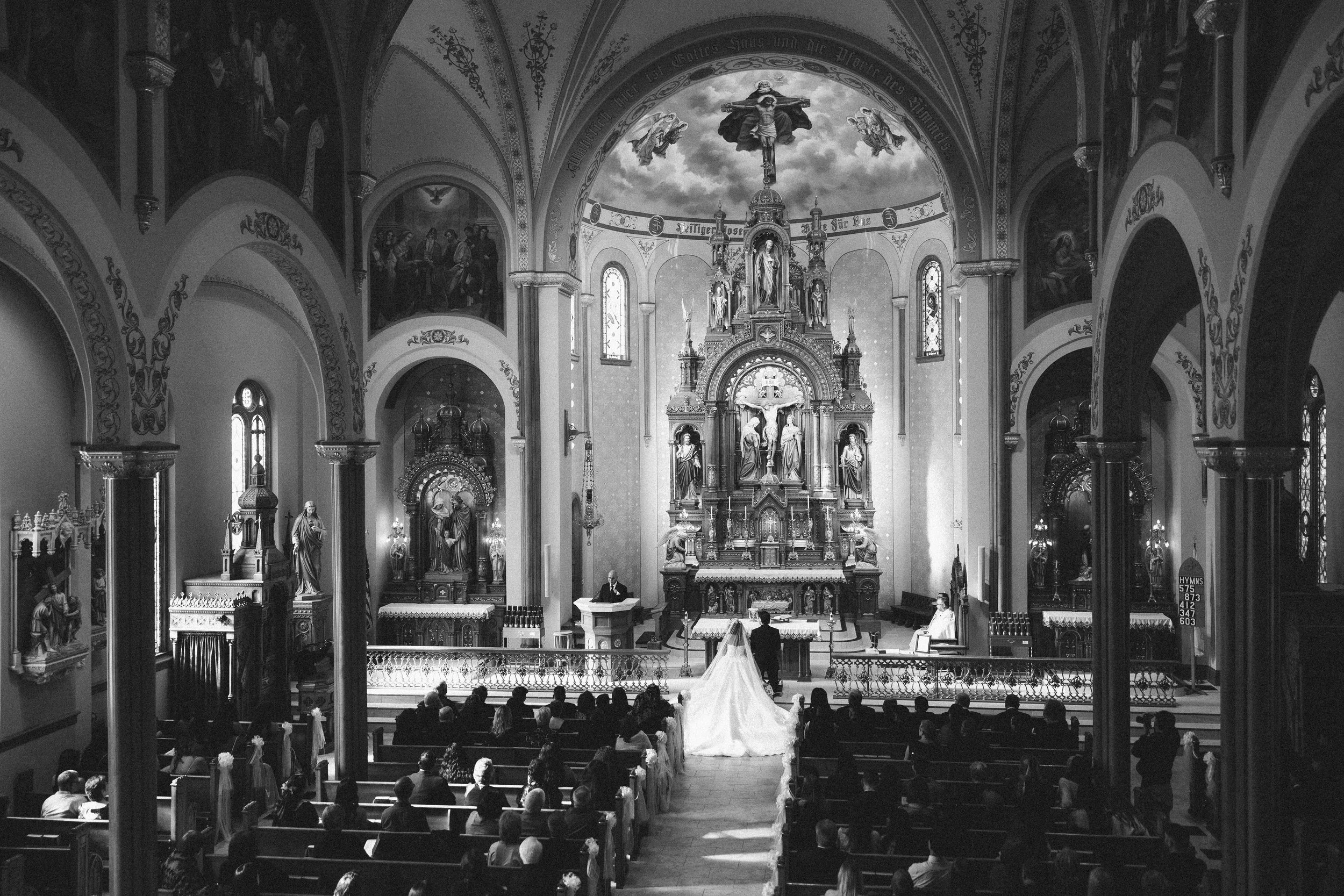 Black and white photo of a church interior during a wedding ceremony, with a bride and groom at the altar facing the priest, surrounded by seated guests.