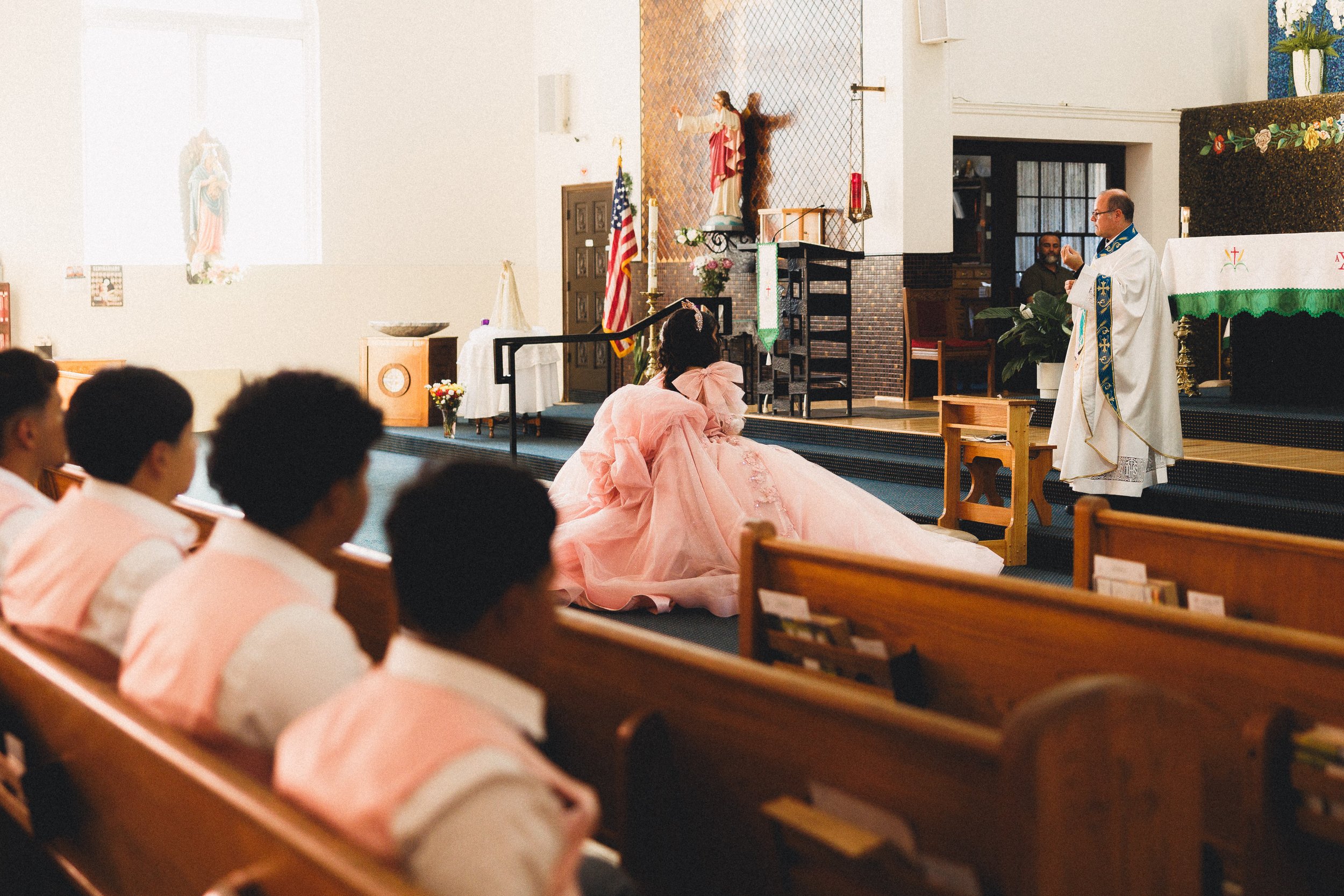 A church wedding ceremony with a bride kneeling before a priest, wearing a pink gown, in front of congregants seated in pews.