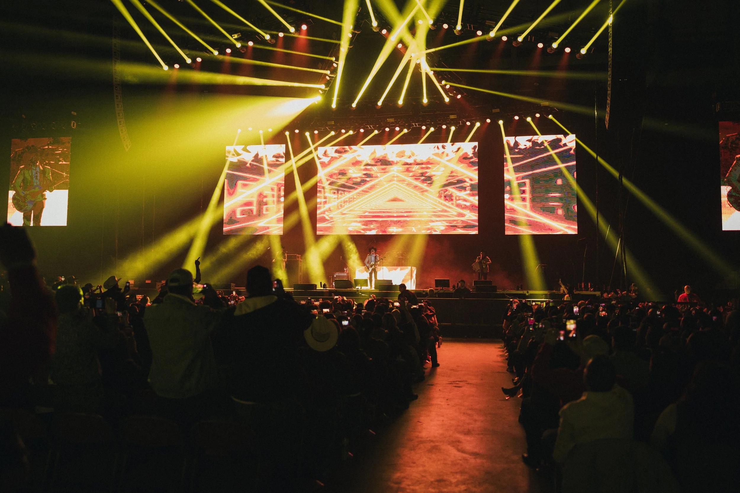 A live concert with a band performing on stage, bright yellow stage lights, large LED screens displaying colorful abstract visuals, and an audience watching and capturing the moment. Los Tucanes de Tijuana Stormont Vail Event Center Topeka, Kansas. 