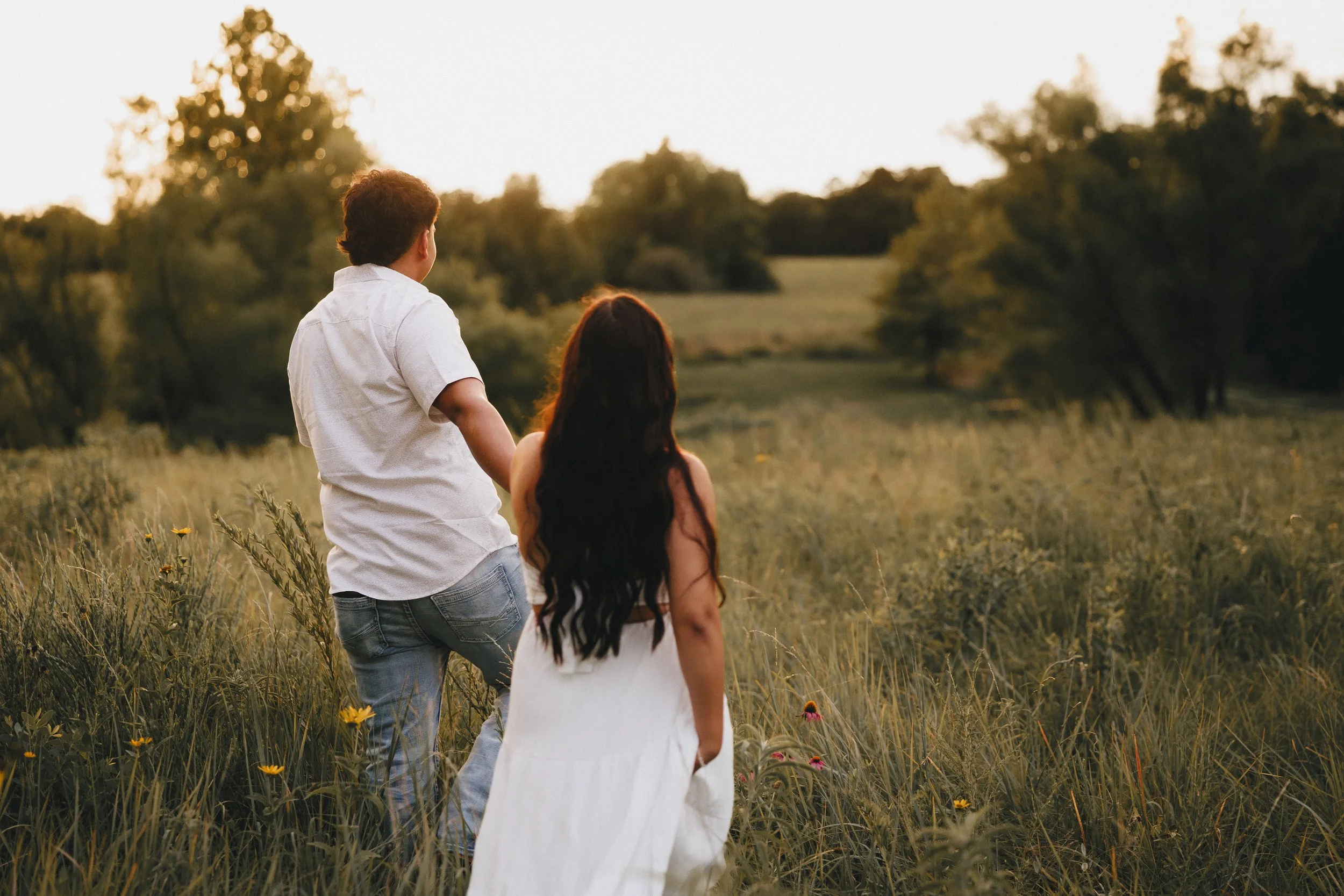 A man and woman holding hands walking through a grassy field at sunset.  Shot in Topeka, Kansas. 