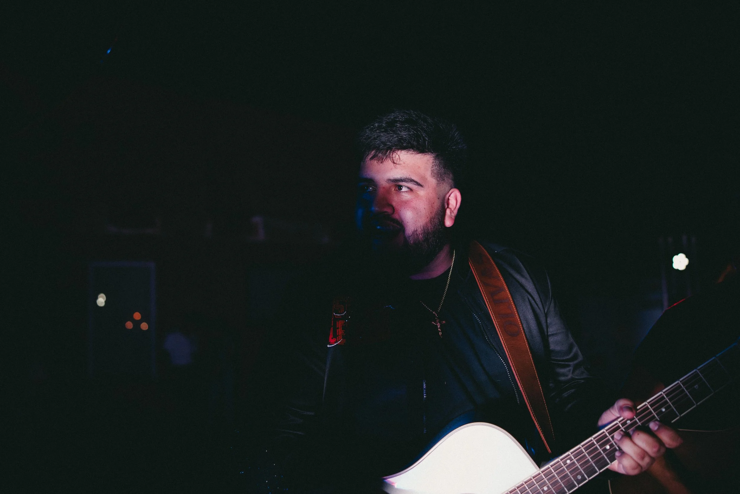 A man with a beard playing a white acoustic guitar in a dark setting, wearing a black leather jacket and a gold necklace. Shot at Fiesta Topeka.