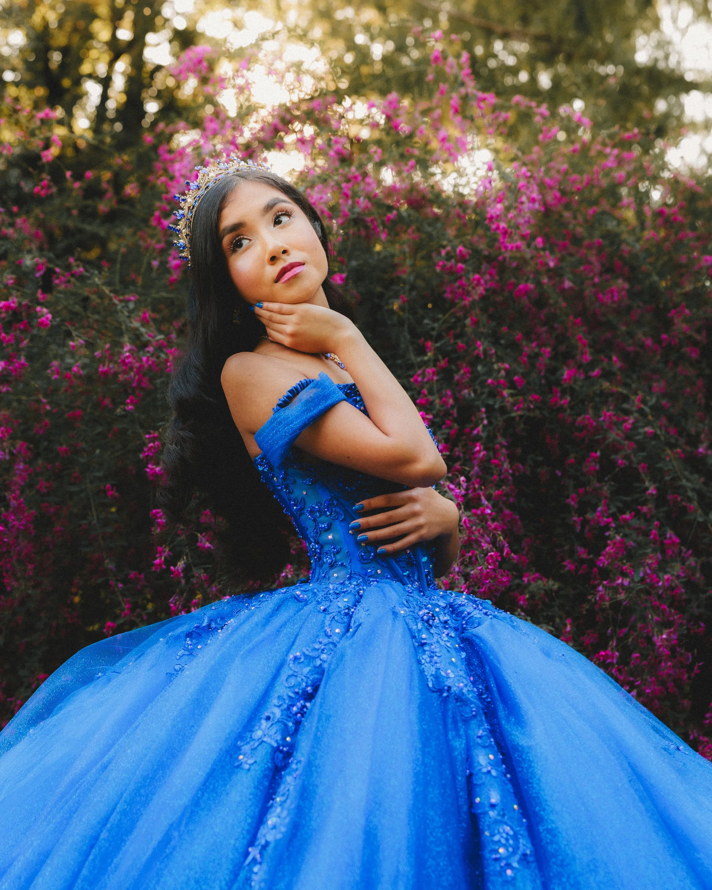 A woman in a blue gown with a tiara, posing outdoors with pink flowers in the background.