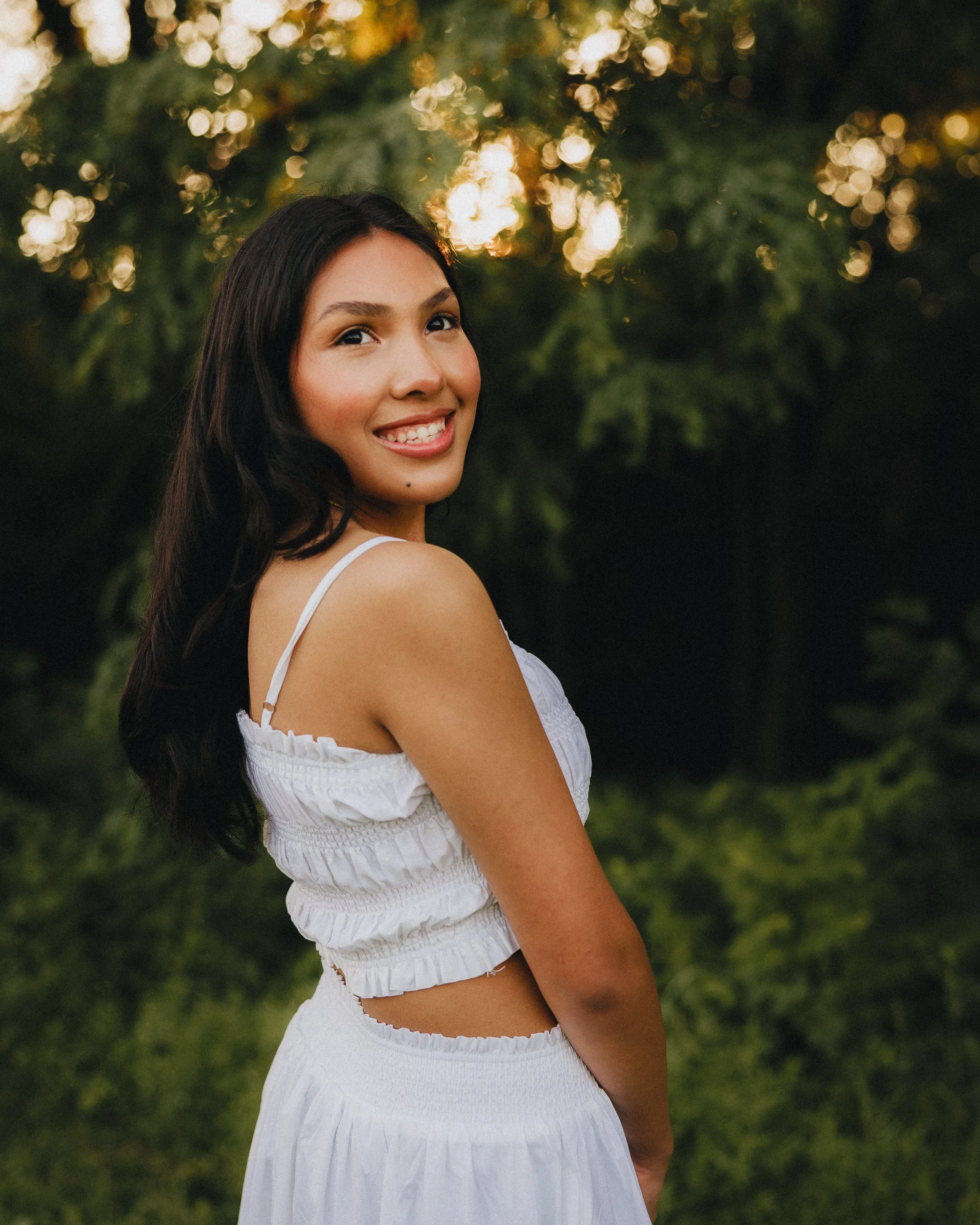 A young woman with long dark hair smiling outdoors at sunset, wearing a white sleeveless top and matching skirt, standing in a green natural setting with blurred trees and sunlight in the background.