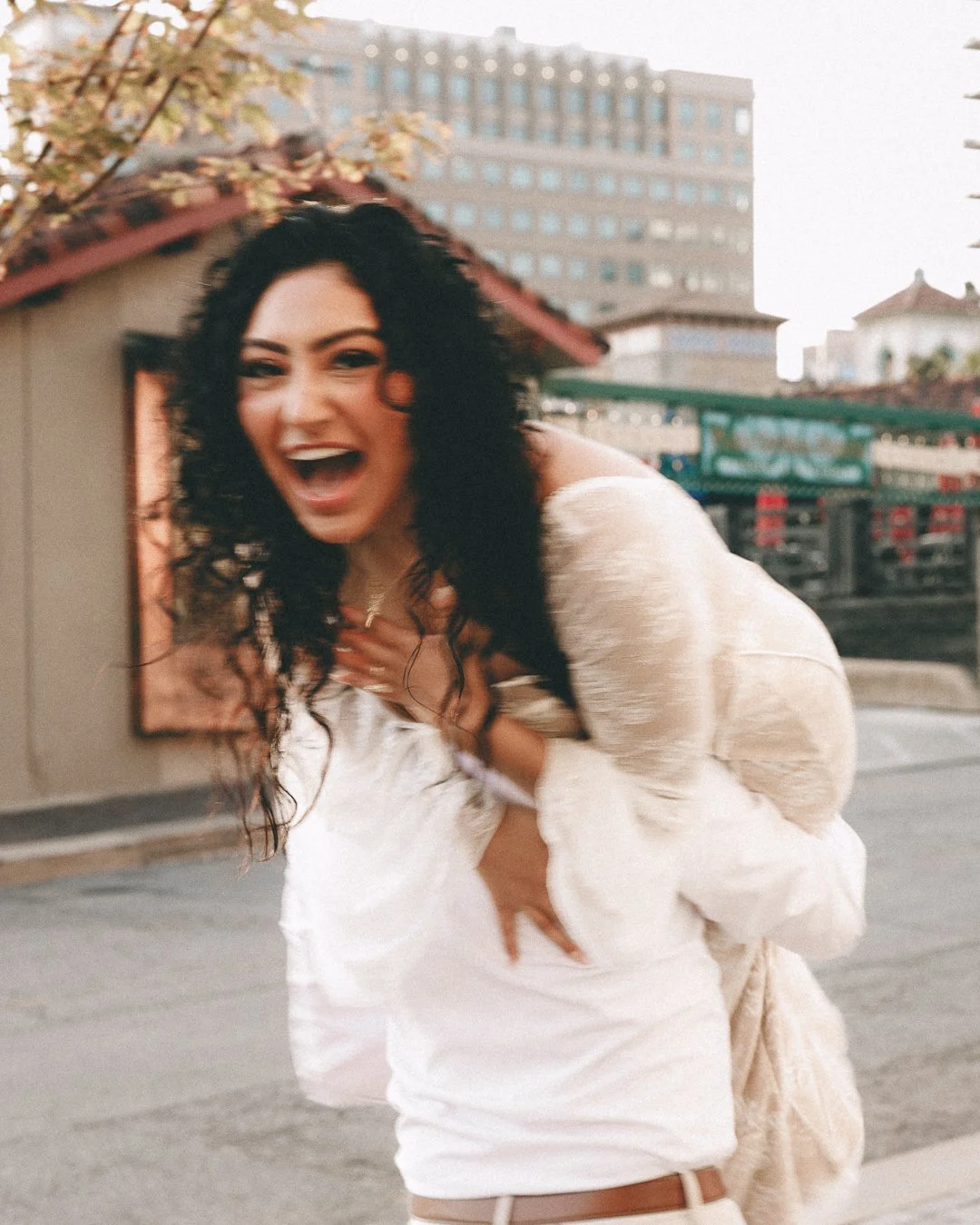 A woman with dark curly hair laughing, holding her chest with one hand and her stomach with the other, outdoors in an urban area with buildings and a tree in the background. Shot at Kansas City, Country Club Plaza.