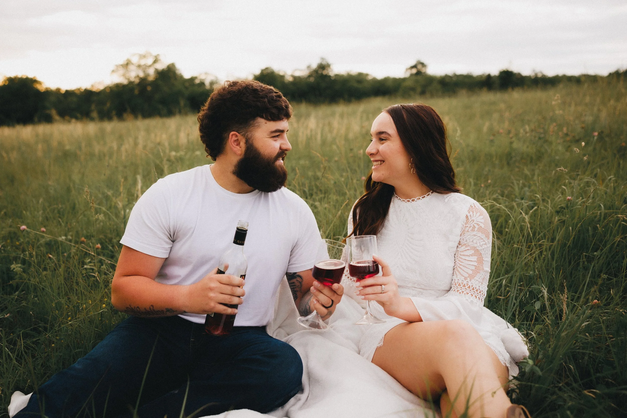 A man and woman sitting in a grassy field having drinks and smiling at each other.