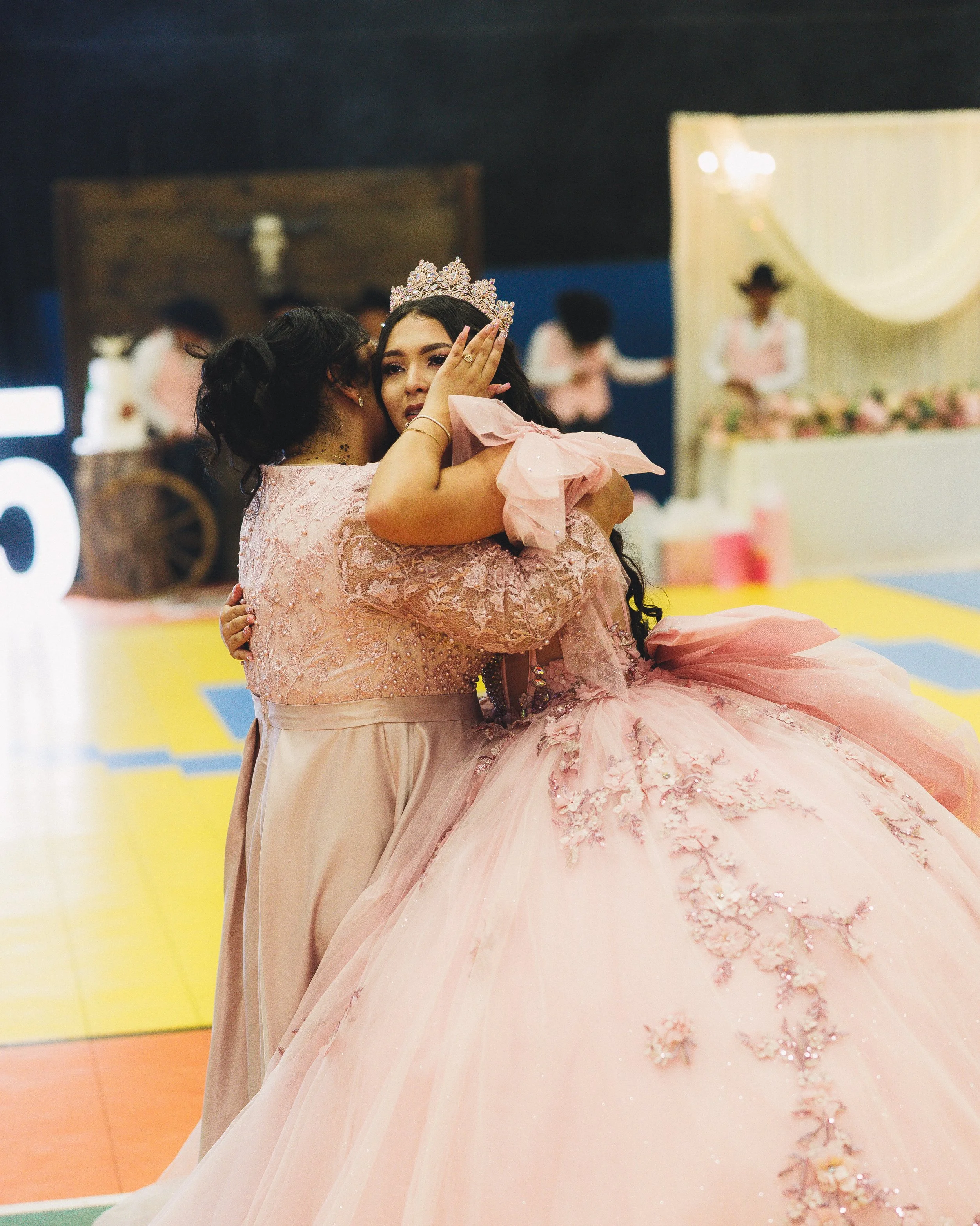A young woman in a pink ball gown and tiara is being embraced and comforted by an older woman in a pink lace dress at a celebration or wedding reception.