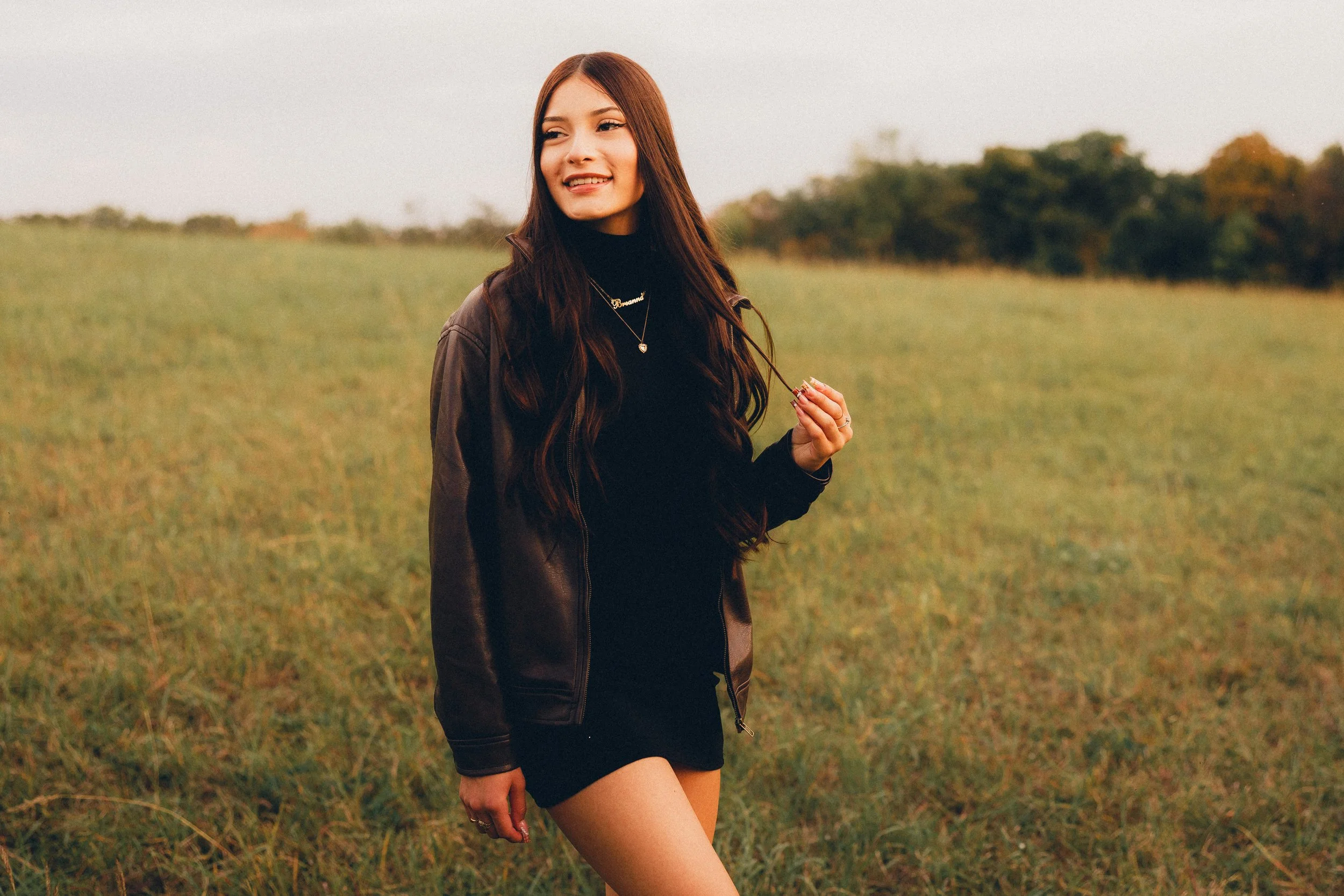 A young woman standing in a grassy field during golden hour, wearing a black dress and leather jacket, holding a necklace and smiling.