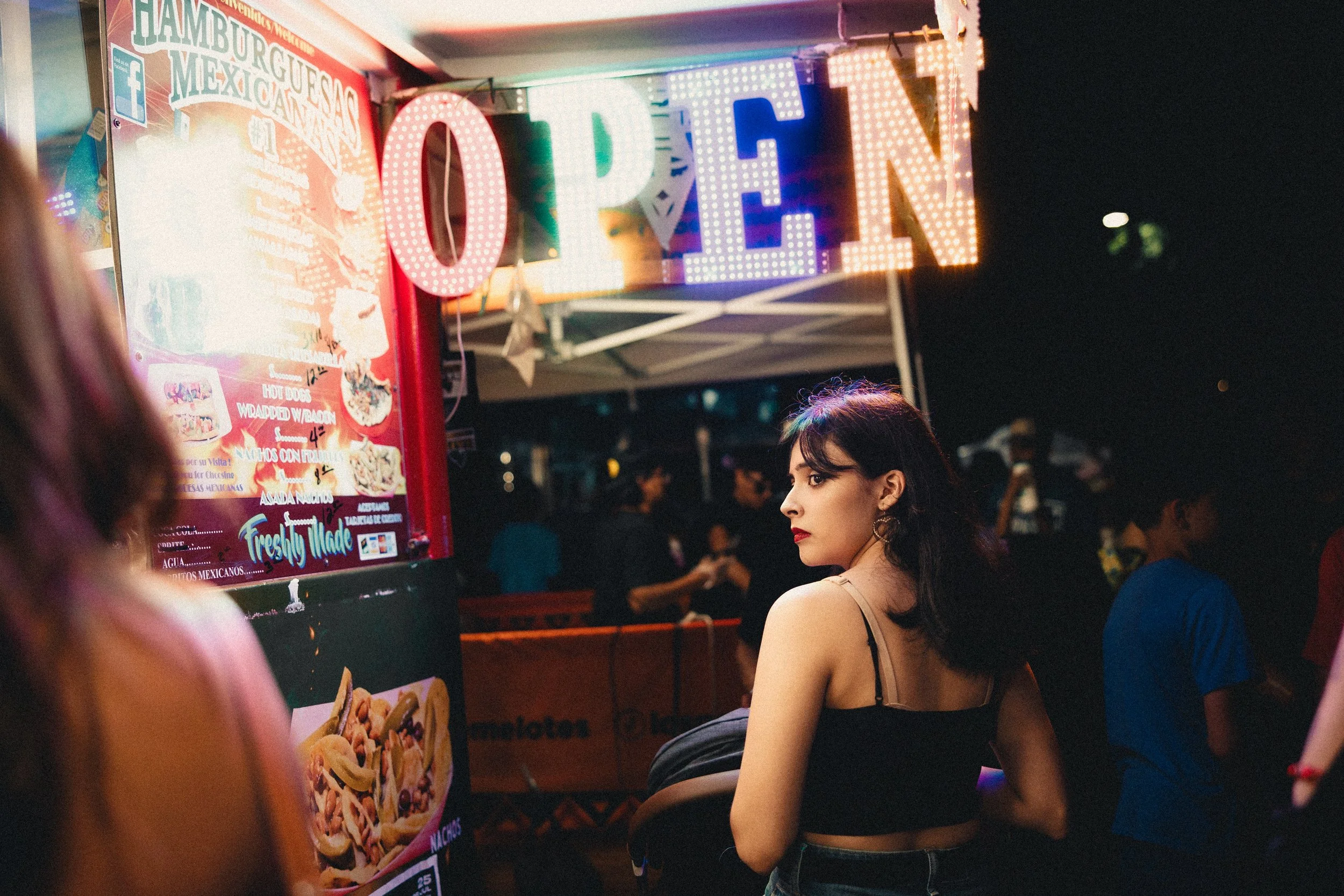 A woman with dark hair and a black top standing in front of a food stand at night, illuminated by bright, colorful lights, with a large illuminated 'OPEN' sign overhead. Shot at Fiesta Topeka.