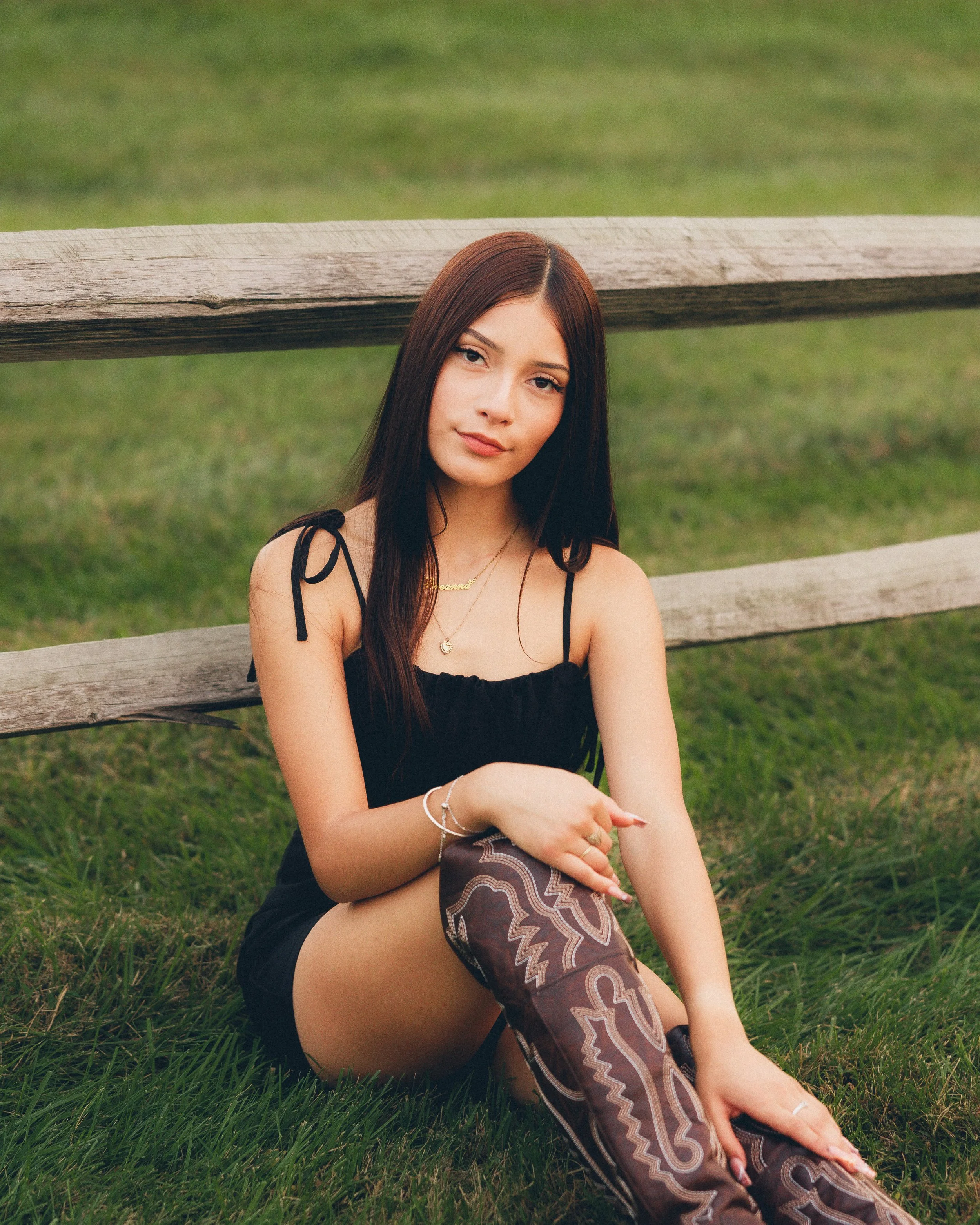 A young woman with long dark hair sitting on grass near a wooden fence wearing a black dress and patterned over-the-knee boots.