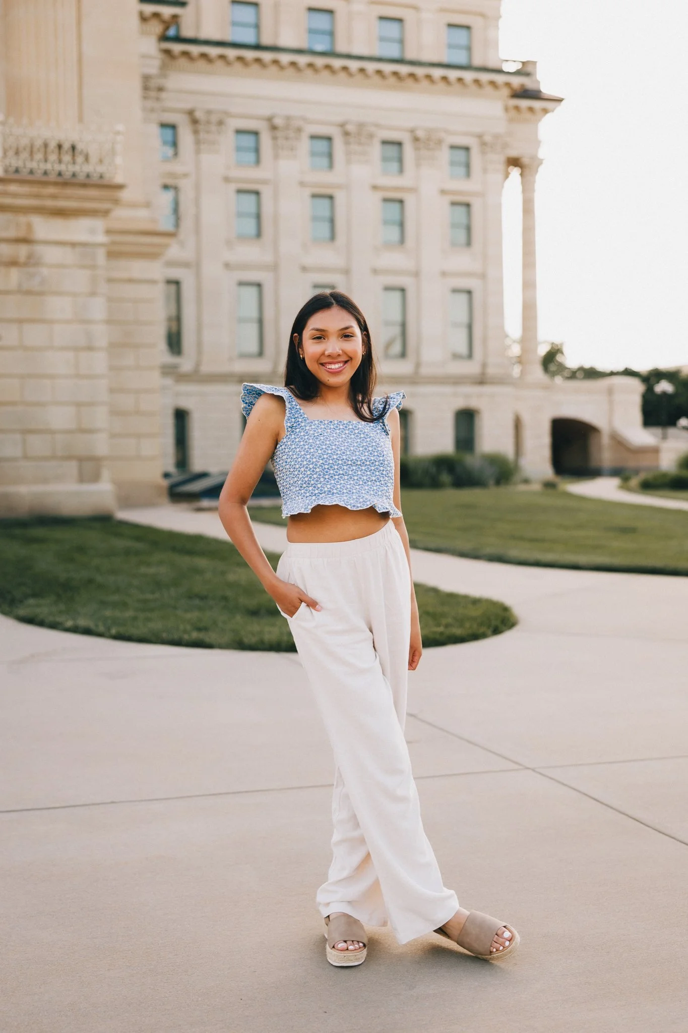 A young woman with dark hair wearing a blue floral crop top and white loose pants standing outdoors in front of a historic building, smiling at the camera. Shot in Topeka, Kansas. 