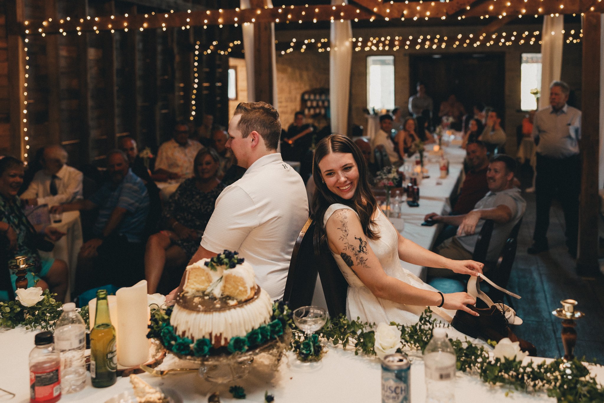 A woman with long dark hair and tattoos on her arm is sitting at a table, smiling and looking back over her shoulder at a wedding reception. She is wearing a white dress and holding a shoe in her hand. There is a man with short hair sitting close by,