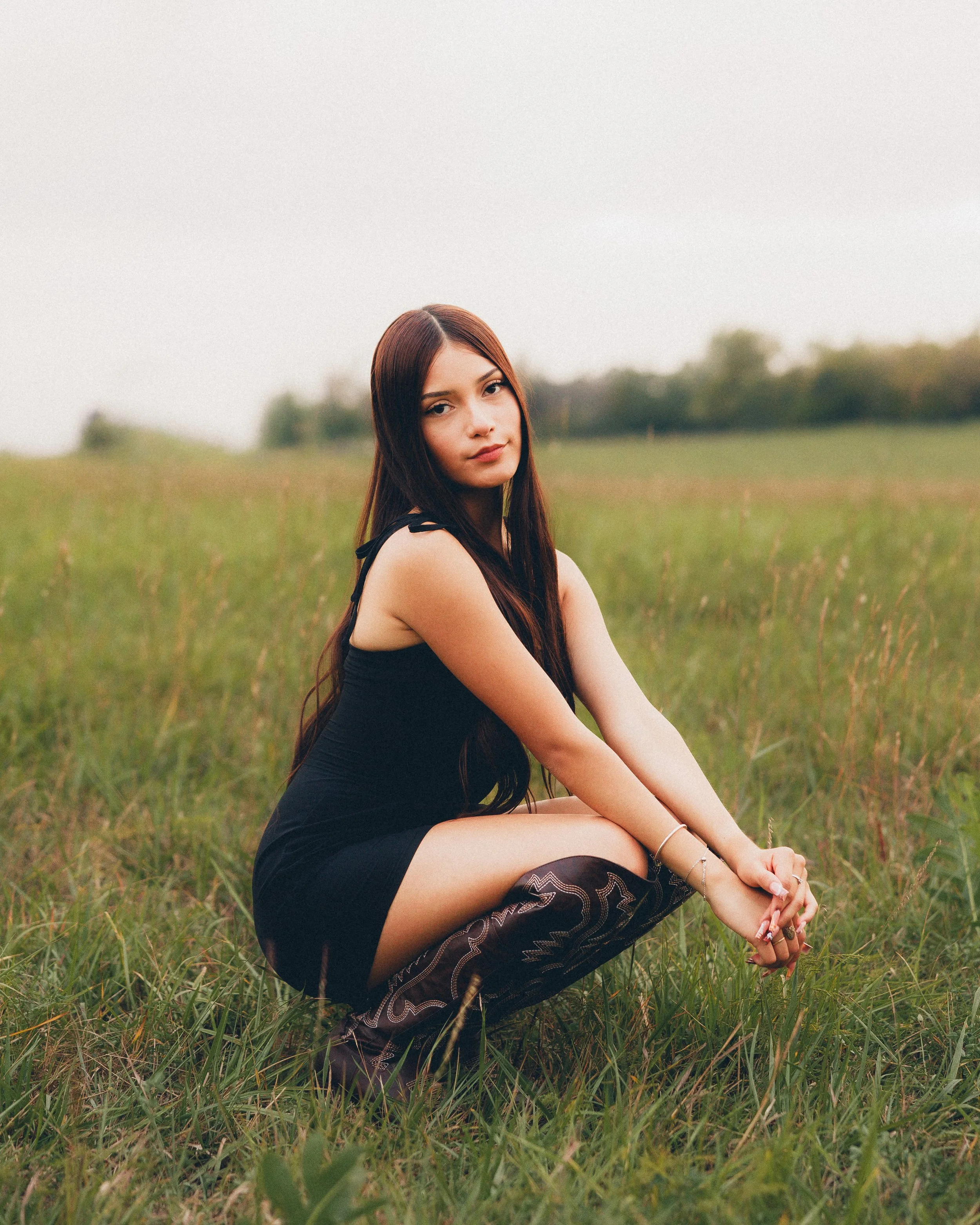 A young woman with long dark hair wearing a black dress and patterned knee-high boots sitting on grass in a field with a blurred natural background.