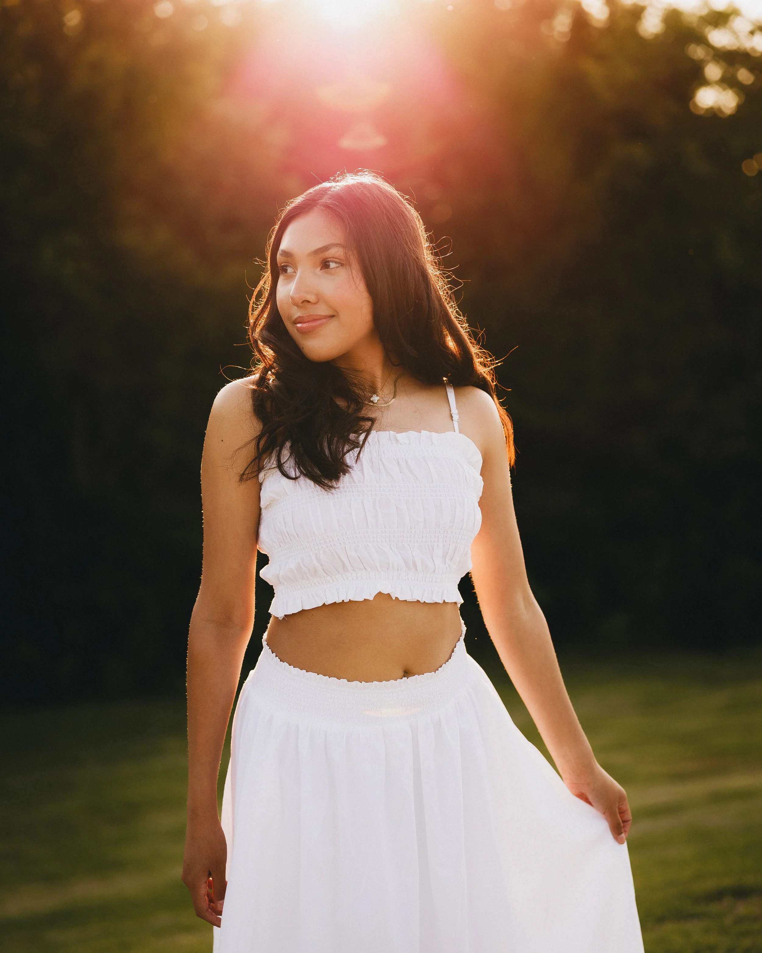 Young woman in white outfit standing outdoors during sunset, with sunlight behind her and trees in the background.
