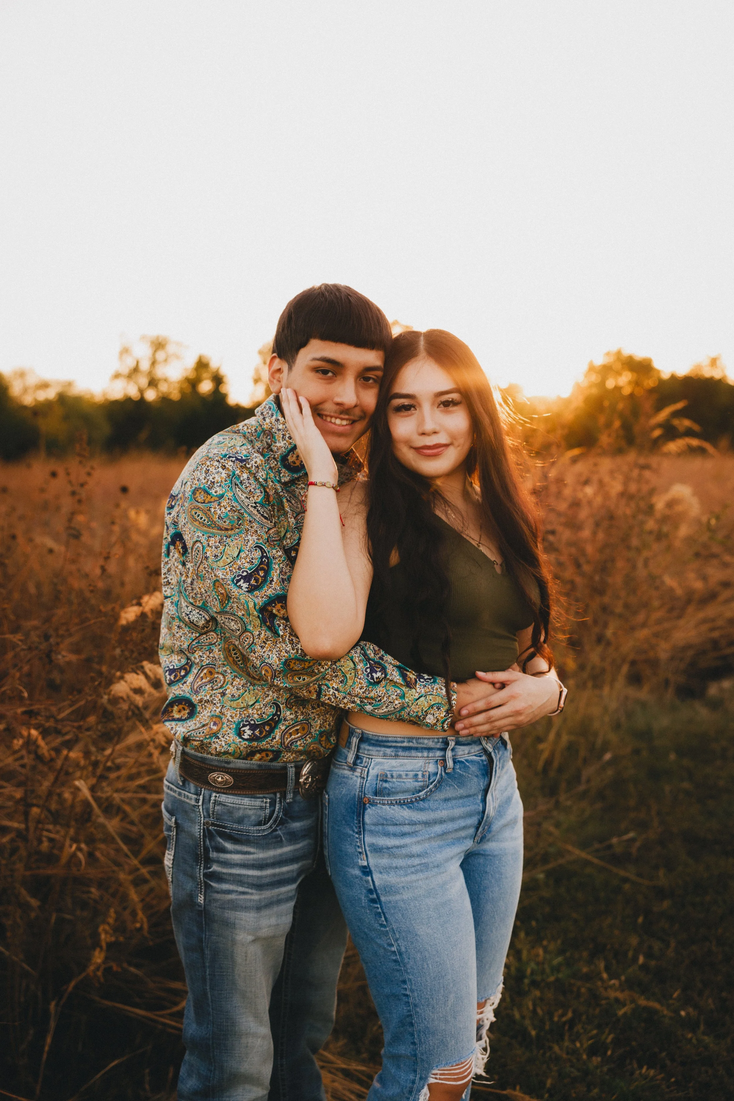 A young man and woman standing close together outdoors during sunset, with the man hugging the woman from behind and both smiling at the camera. Shot in Topeka, Kansas. 