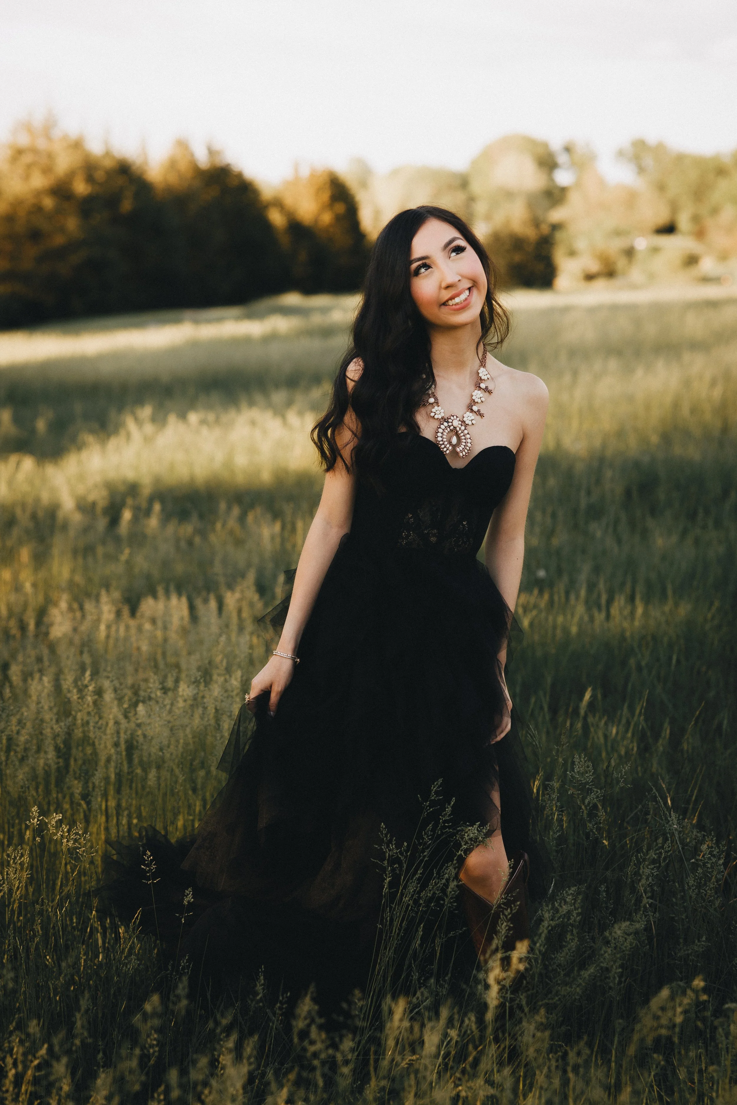 A woman in a strapless black dress and jewelry standing in a grassy field during sunset.