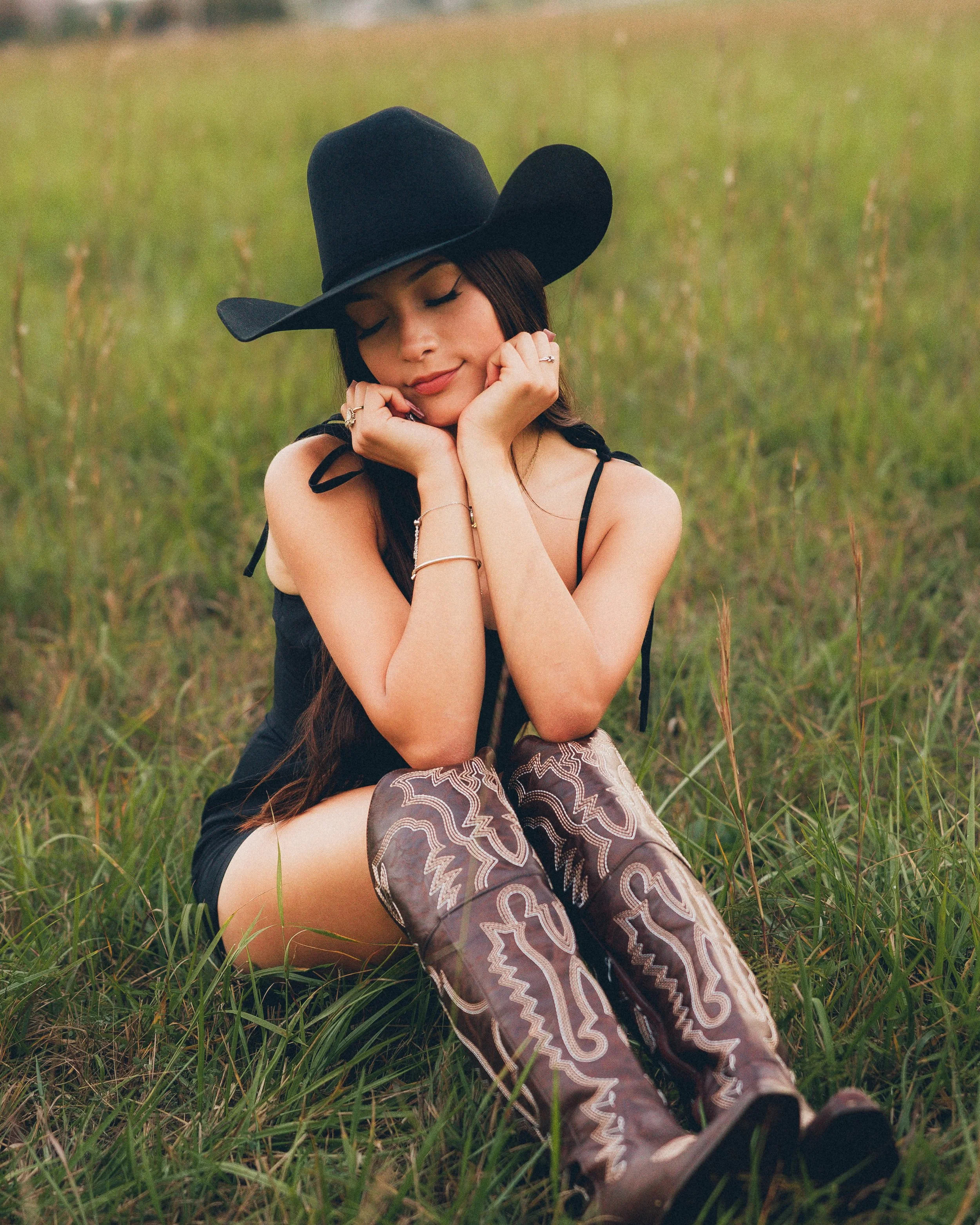 A woman sitting in a grassy field, wearing a large black hat and patterned knee-high boots, with her eyes closed and hands resting on her cheeks.