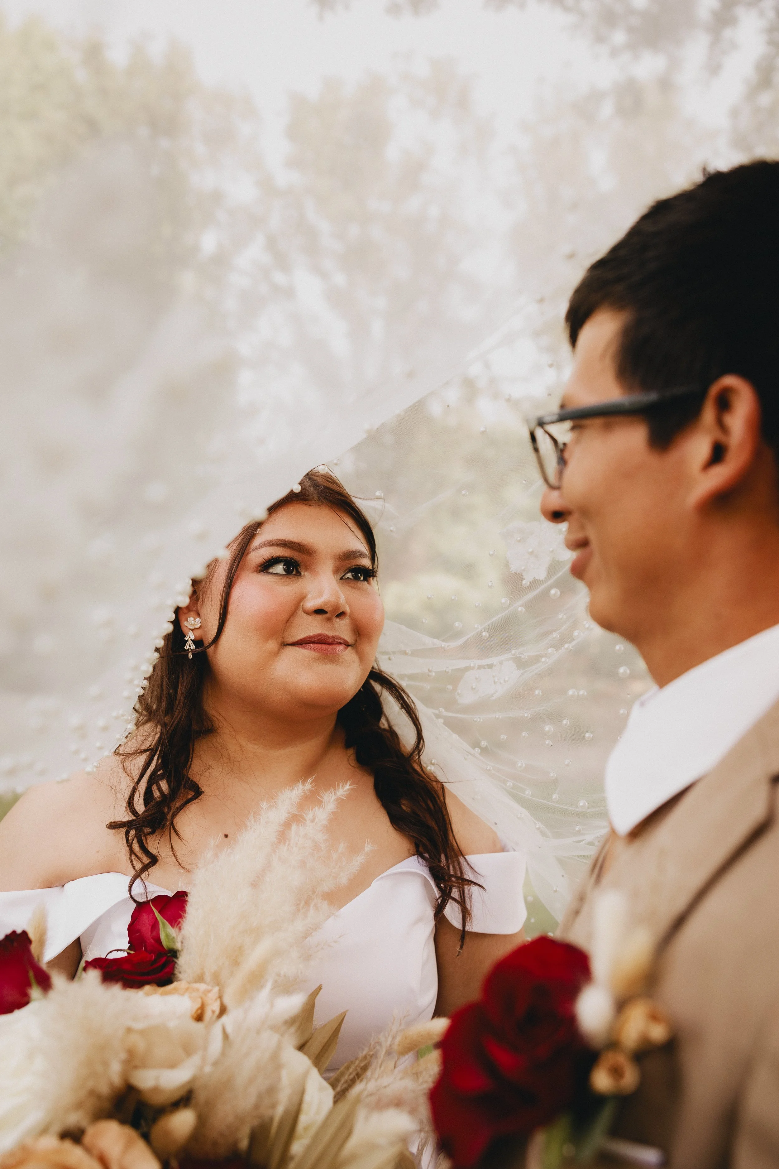 A bride and groom facing each other outdoors, the bride holding a bouquet of roses and dried flowers, with the bride looking at the groom under her veil with floral decoration.